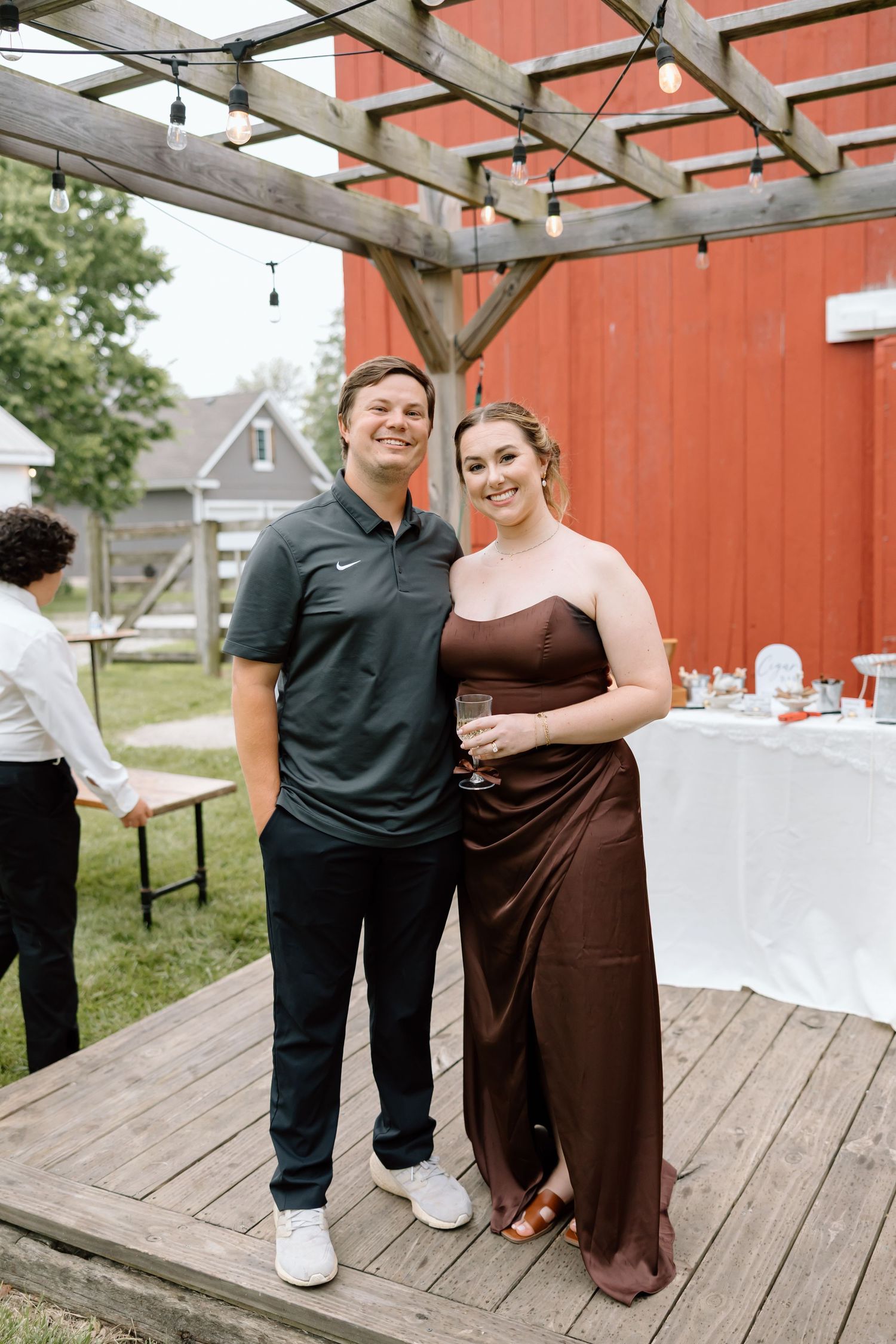 A couple poses outdoors at a rustic barn wedding reception, man in Nike polo and woman in brown satin gown.