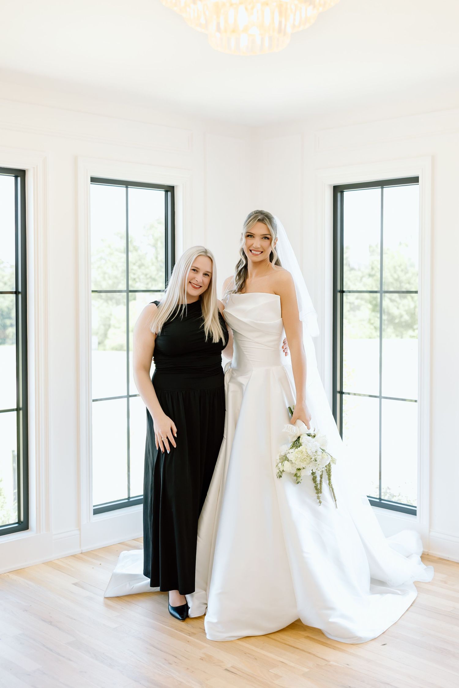 Bride in white strapless gown holding bouquet poses with woman in black dress in bright white room.