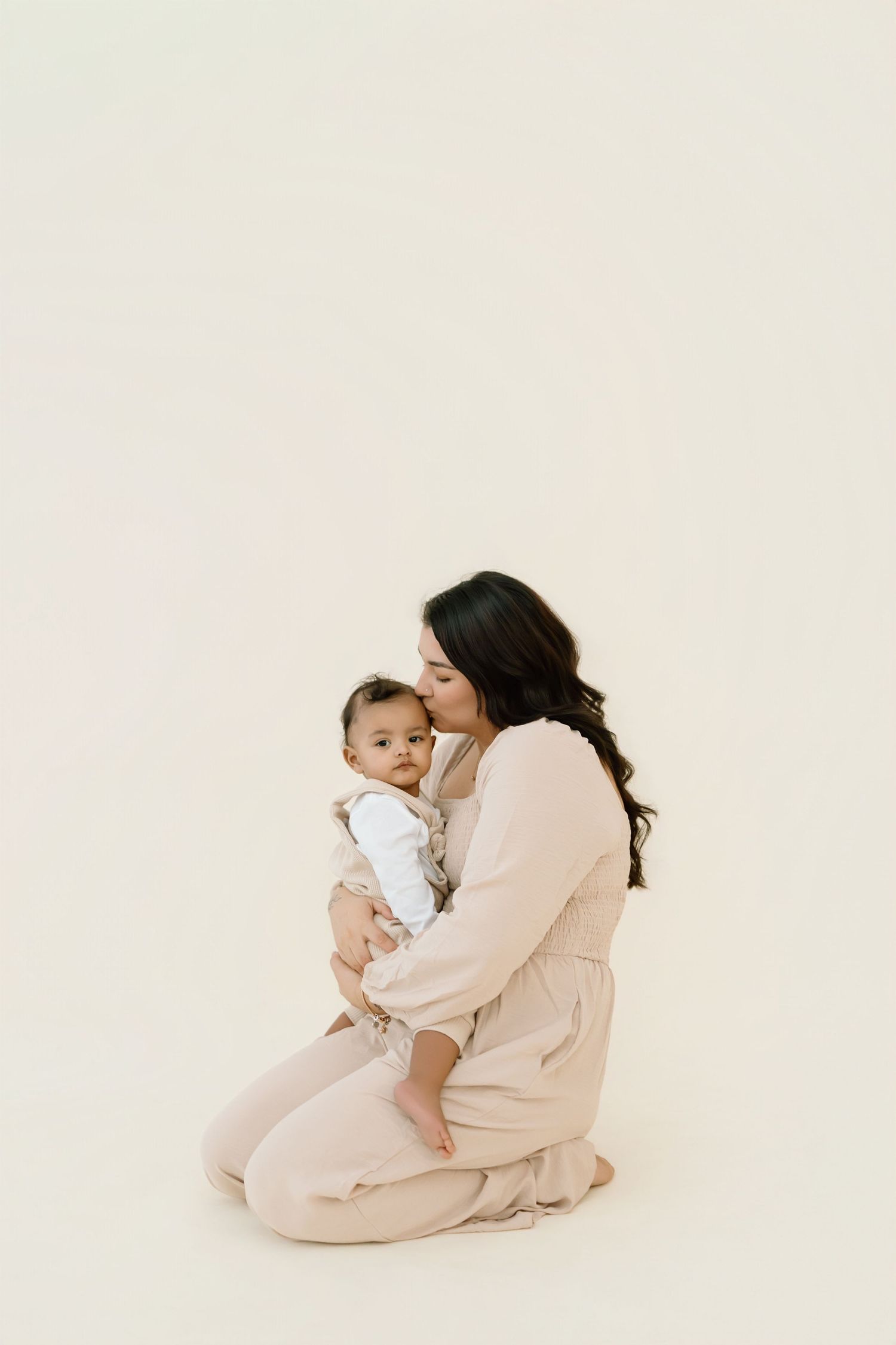 A tender moment captured in soft neutral tones as parent and baby share loving interactions in minimalist studio setting.