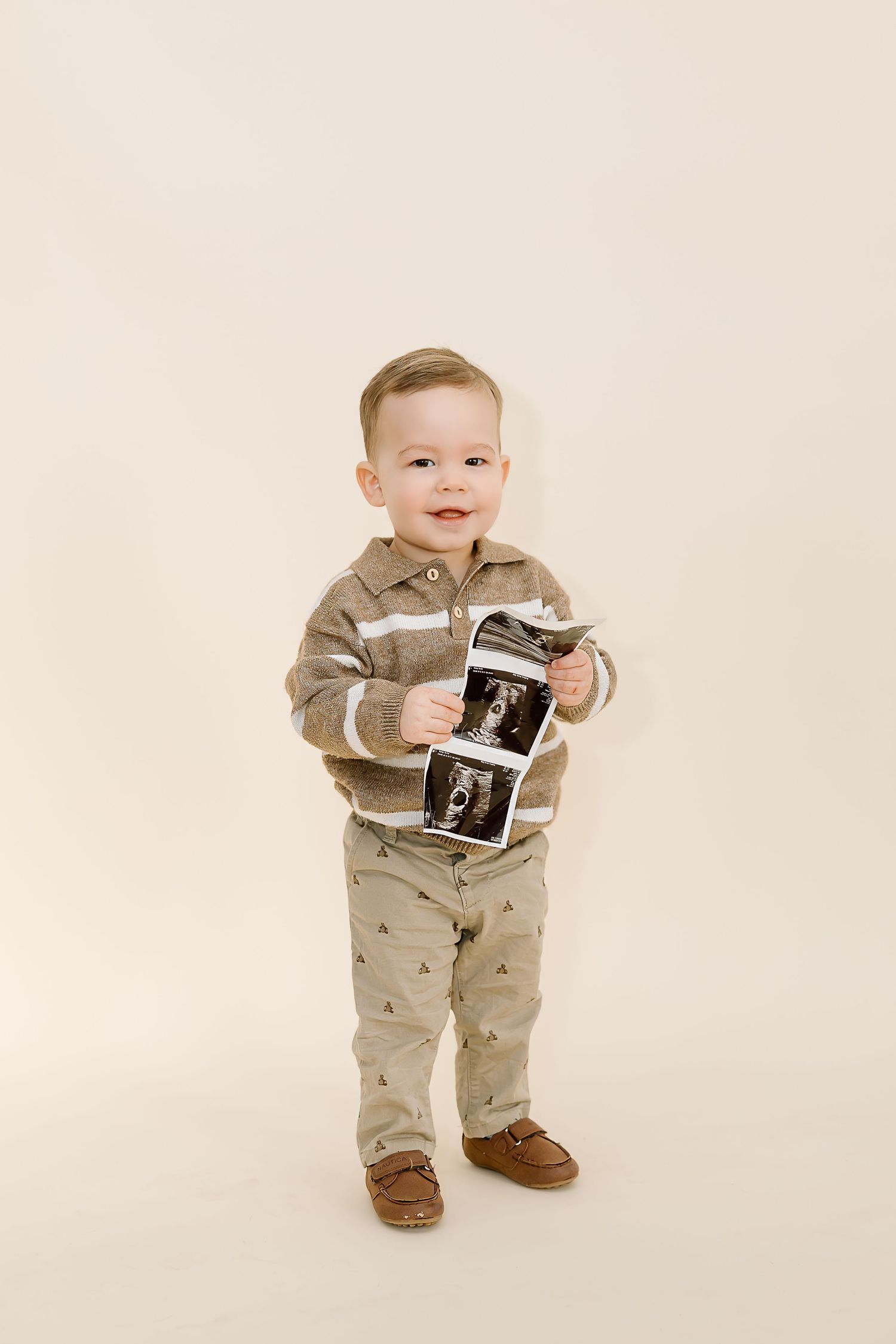 A small child in a tan outfit holds a black and white ultrasound photo while standing against a white backdrop.