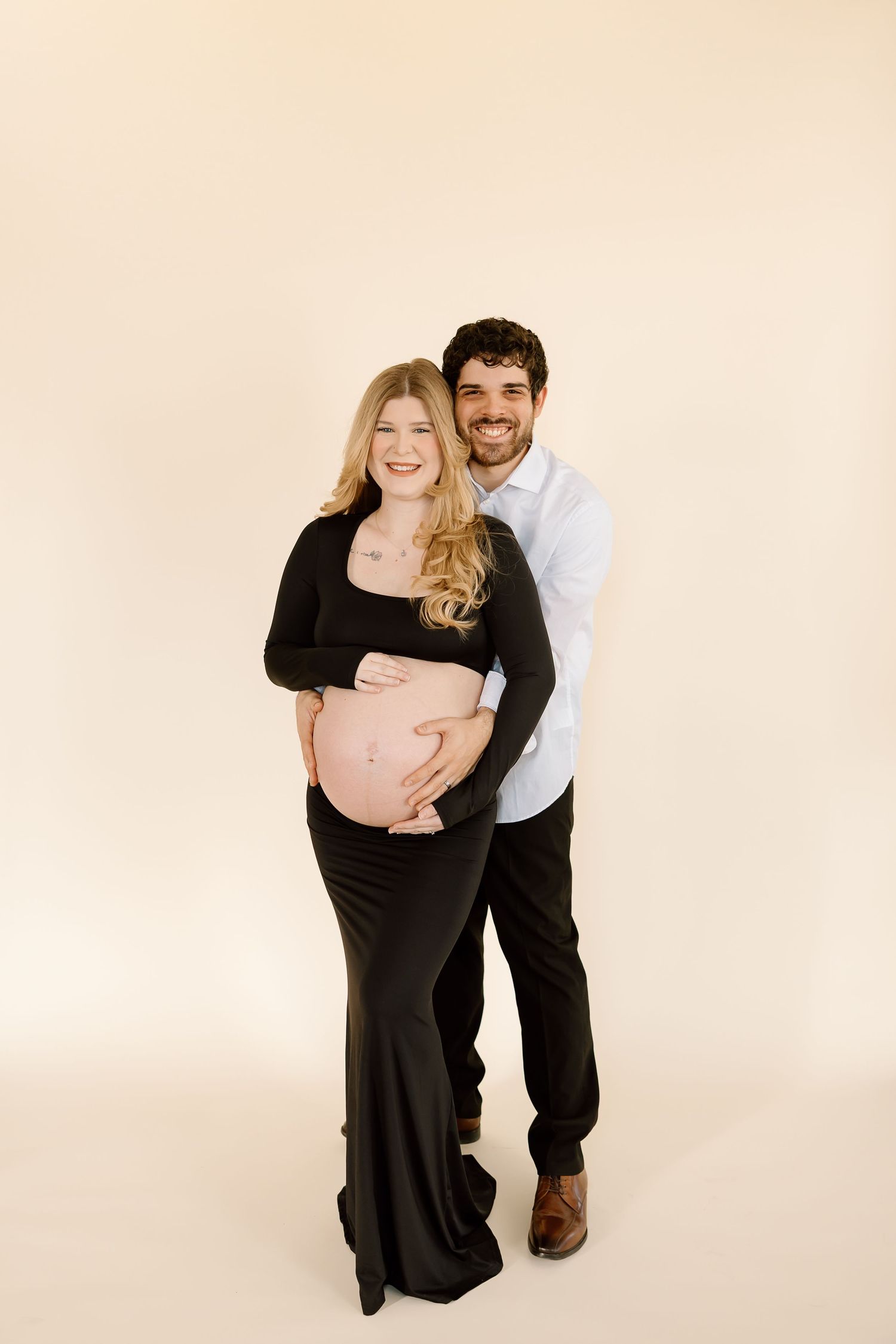 A couple poses together for a maternity photo against a cream background, with the woman wearing a black dress.