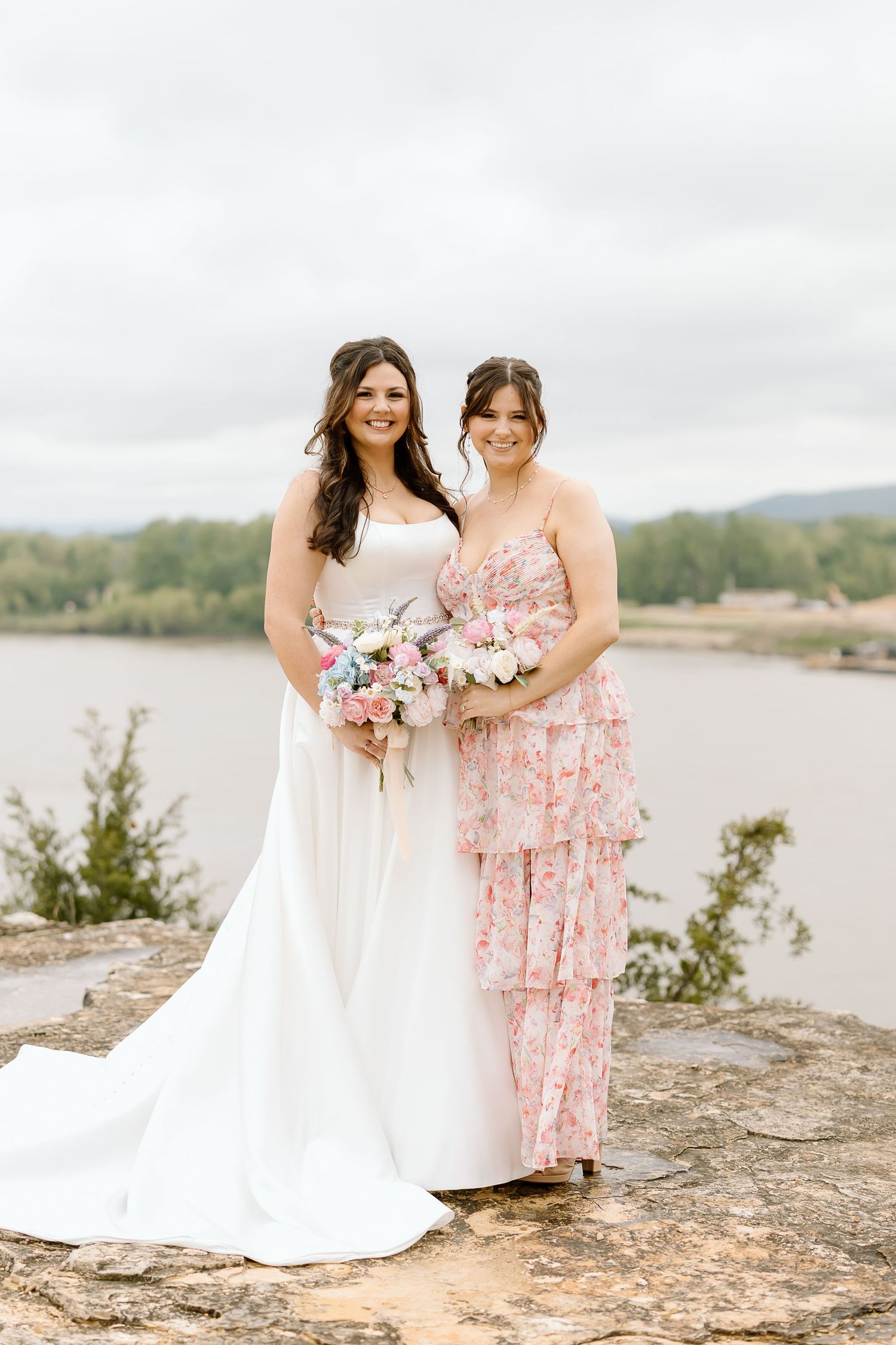 Two people in white and pink floral dresses stand together on rocky terrain near a scenic waterfront.
