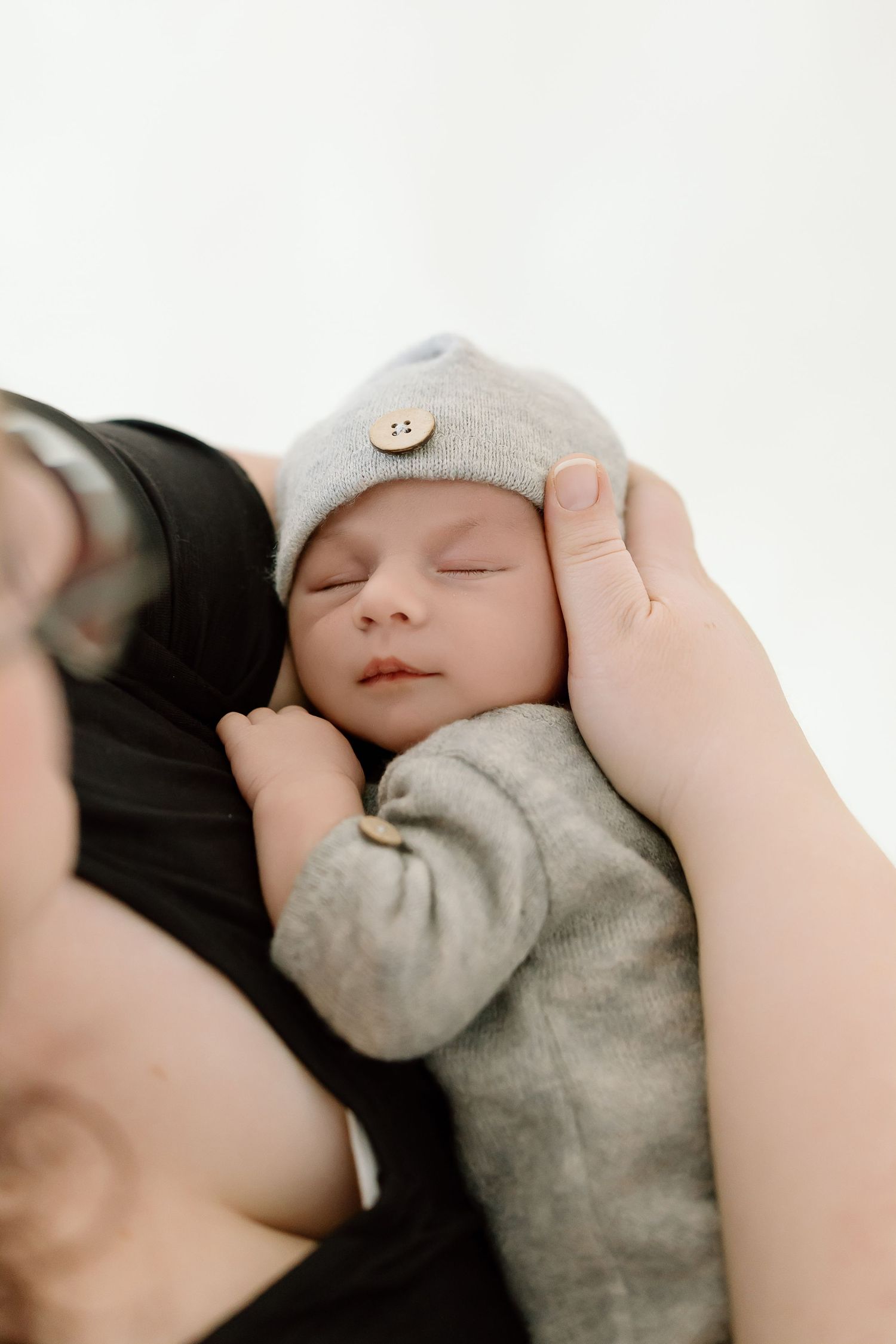 Sleeping newborn baby wearing a soft gray knit hat being gently cradled by adult hands.