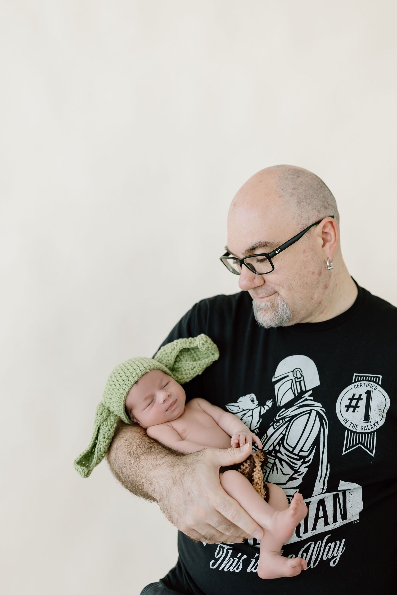 A sleeping newborn wearing a green knit hat is cradled in the arms of someone wearing a black graphic t-shirt.