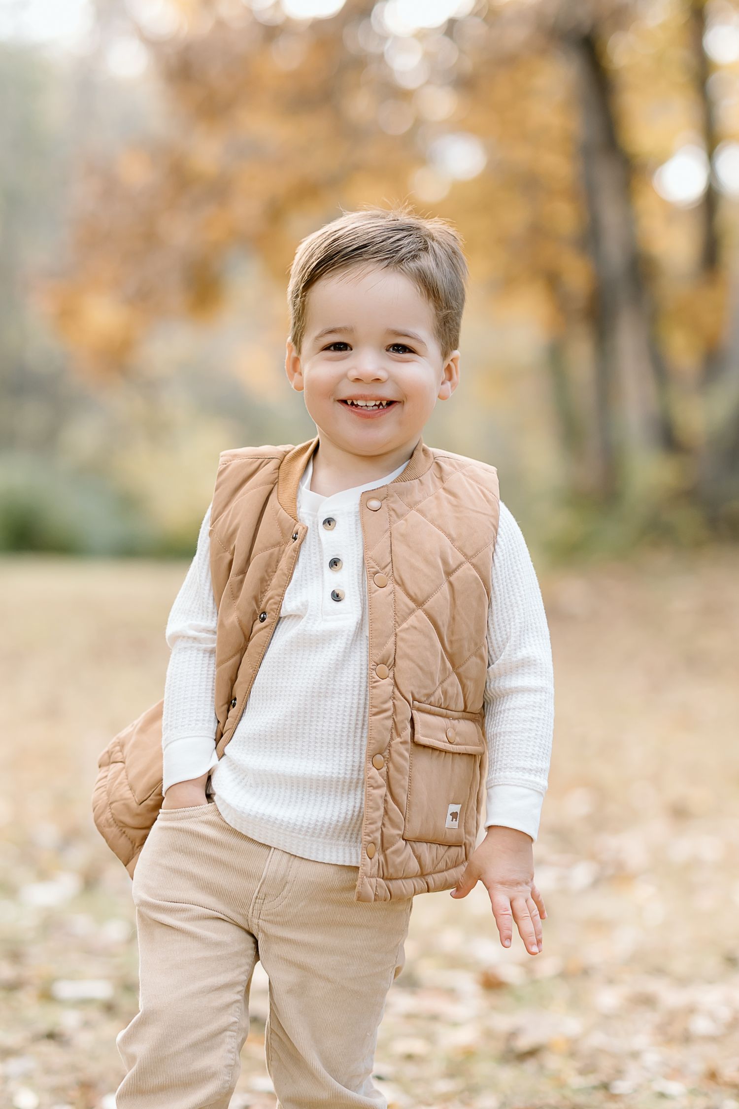 Child wearing brown quilted vest and white sweater plays in autumn leaves during outdoor photo session.