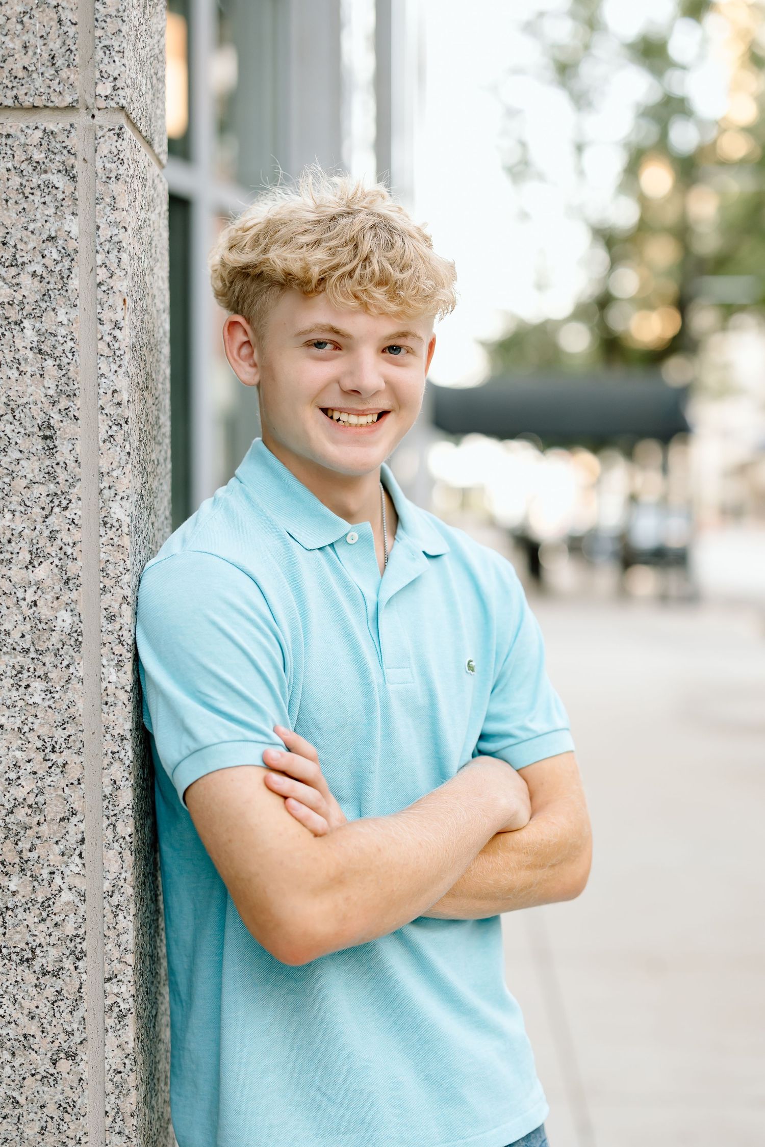A smiling figure in a light blue polo shirt poses against a stone pillar outdoors.
