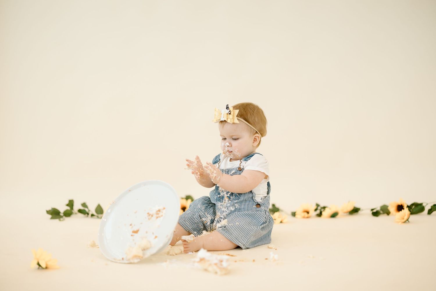 A baby in denim overalls sits and plays with toys on a cream-colored backdrop in a photo series.