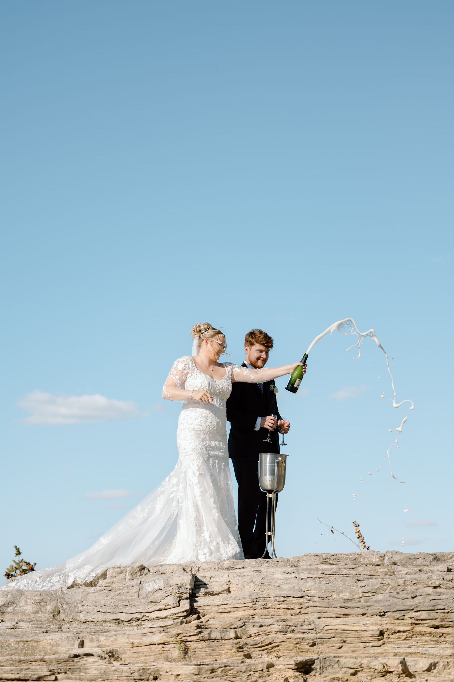 Wedding couple celebrates with champagne on a scenic cliff as confetti flies through the air.