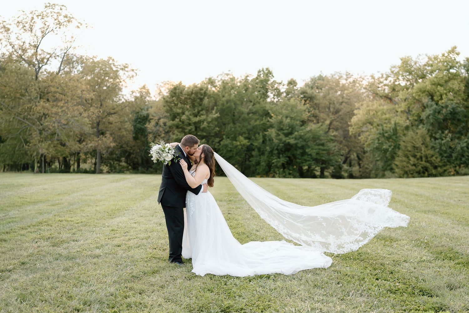 Romantic wedding couple embraces in a sunlit meadow with a flowing white veil catching the breeze.
