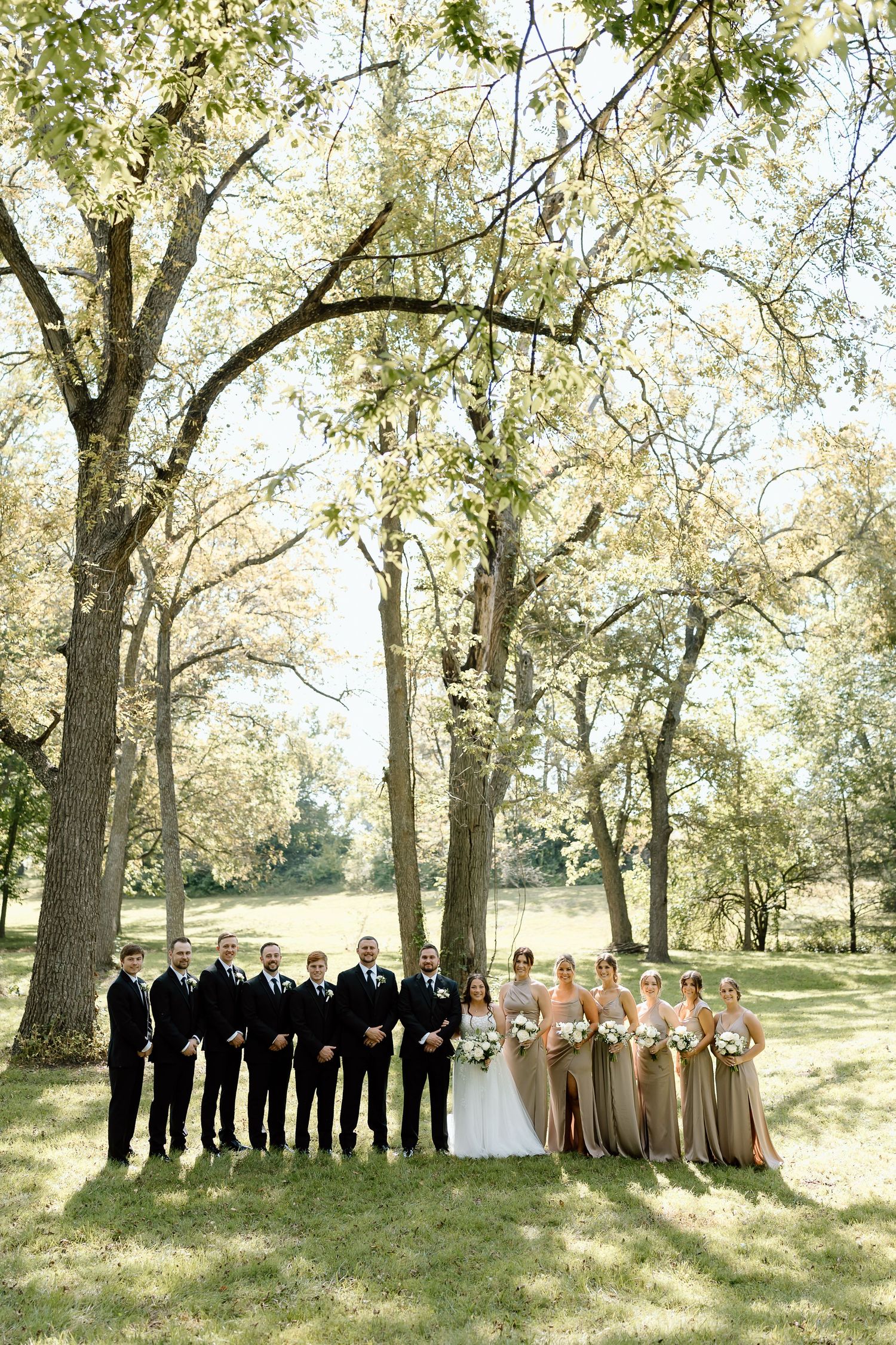 Wedding party stands in a line under tall trees on a sunny day, with groomsmen in black and bridesmaids in neutral gowns.