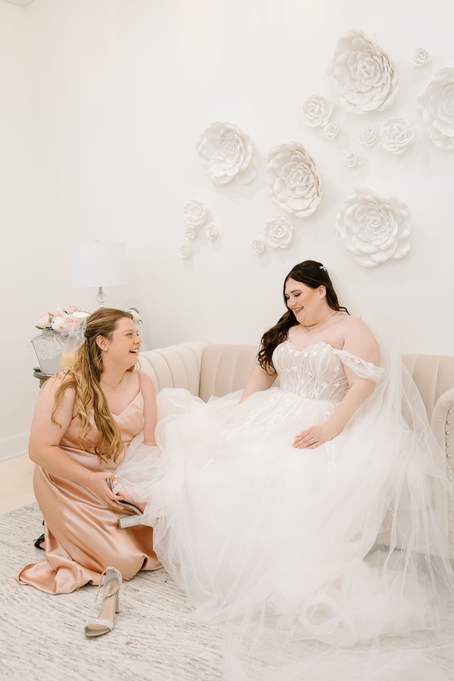 Two people in soft pink and white dresses share joyful moments during a photo session with floral wall decor.