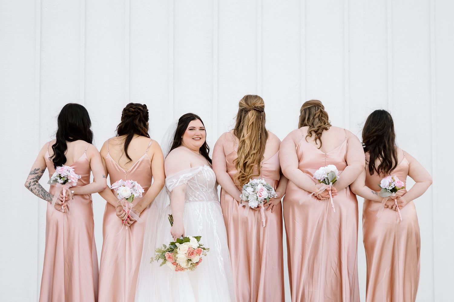 A row of bridesmaids in blush pink dresses stand with their backs to the camera holding white floral bouquets.
