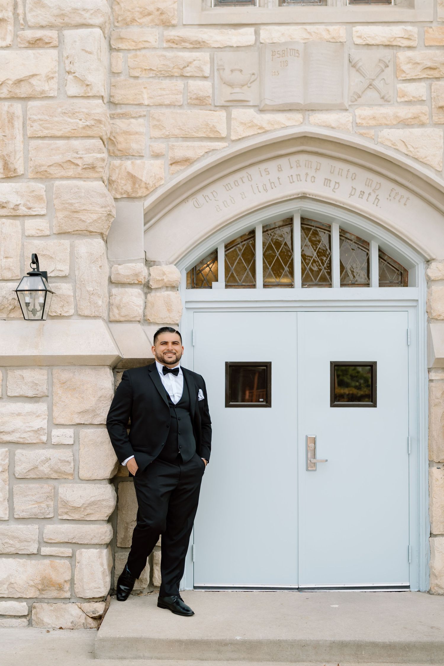 Groom in black tuxedo posing in front of stone church with light blue doors on wedding day.