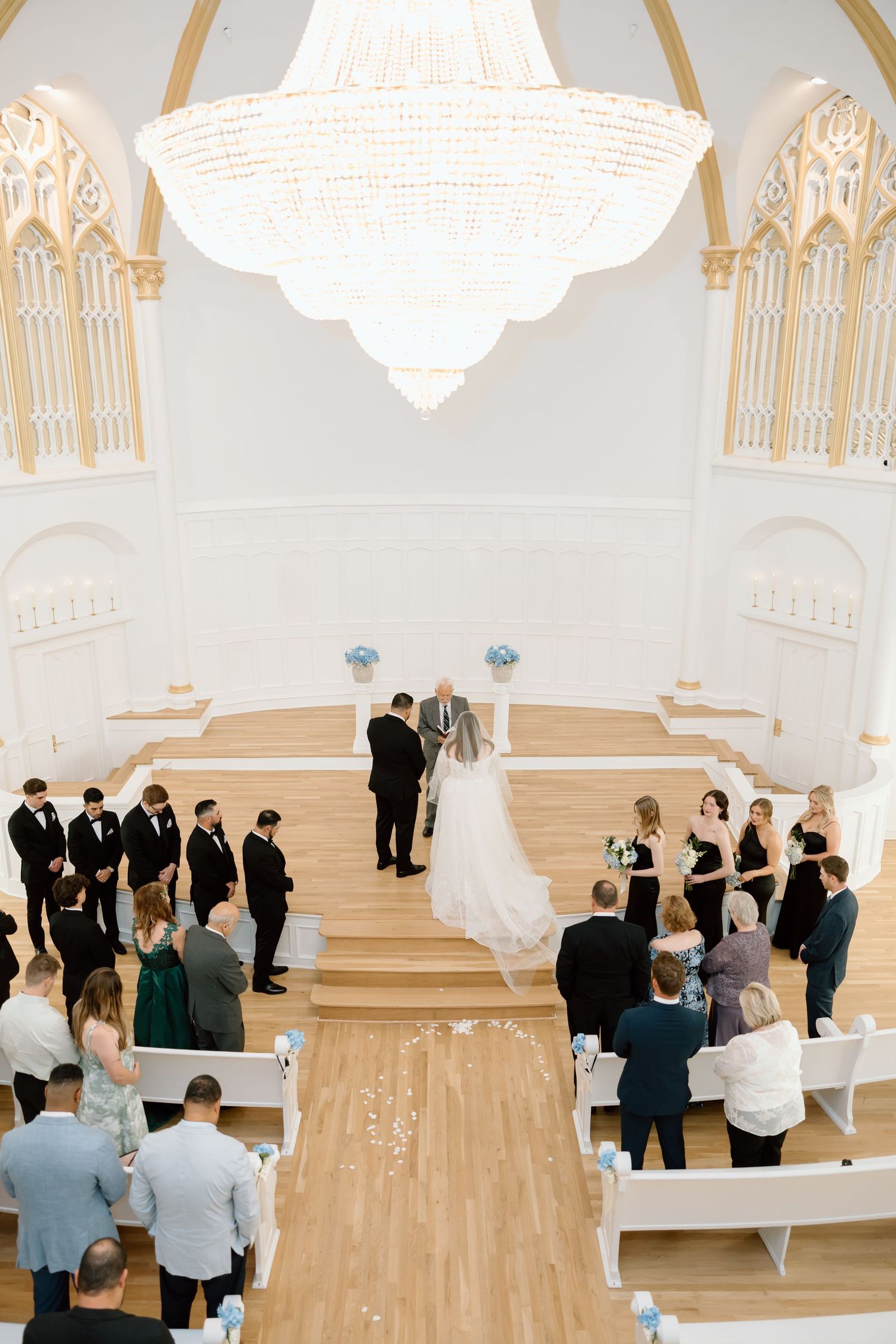 Aerial view of elegant white church wedding ceremony with chandelier, bridal party at altar, and guests in pews.