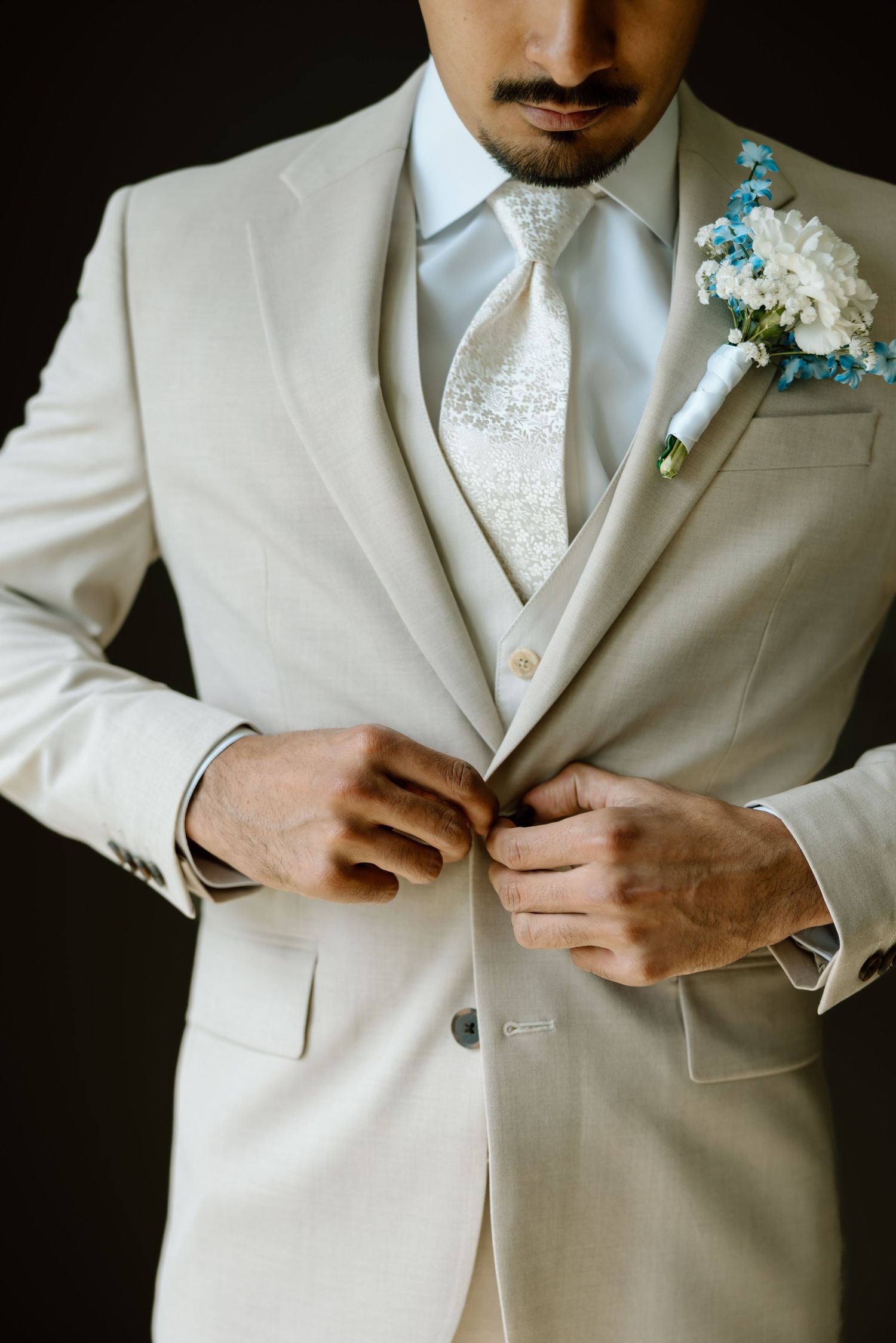 Groom in cream suit with floral boutonniere buttoning jacket before wedding ceremony.