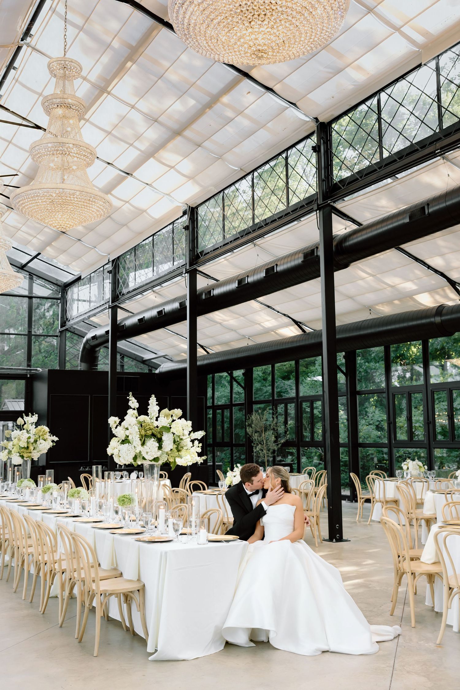Bride and groom share a kiss at elegantly set wedding reception table in a stunning glass greenhouse venue.