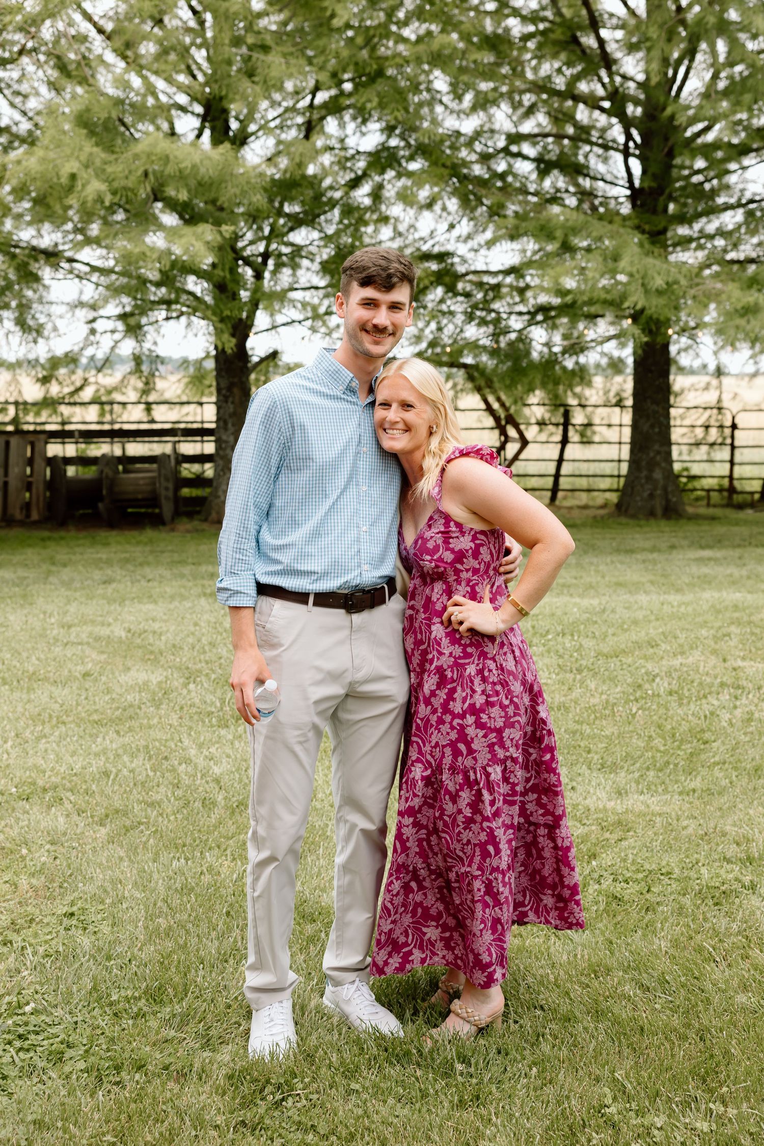 A tall young man and smiling blonde woman pose together in a green field near wooden fencing and large trees.