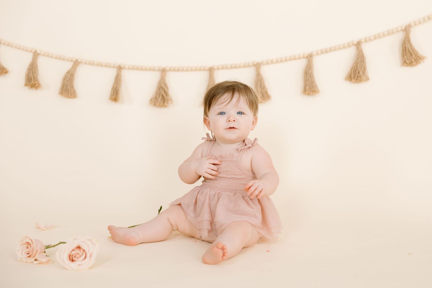 A baby in dusty rose clothing sits with pink roses and a tassel banner decoration.