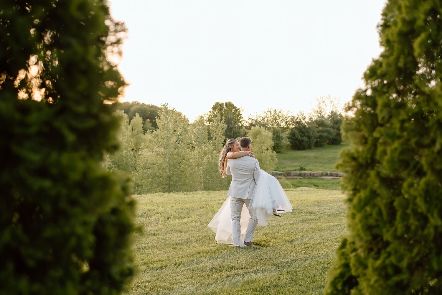 Groom carries bride through garden path during sunset wedding photos.
