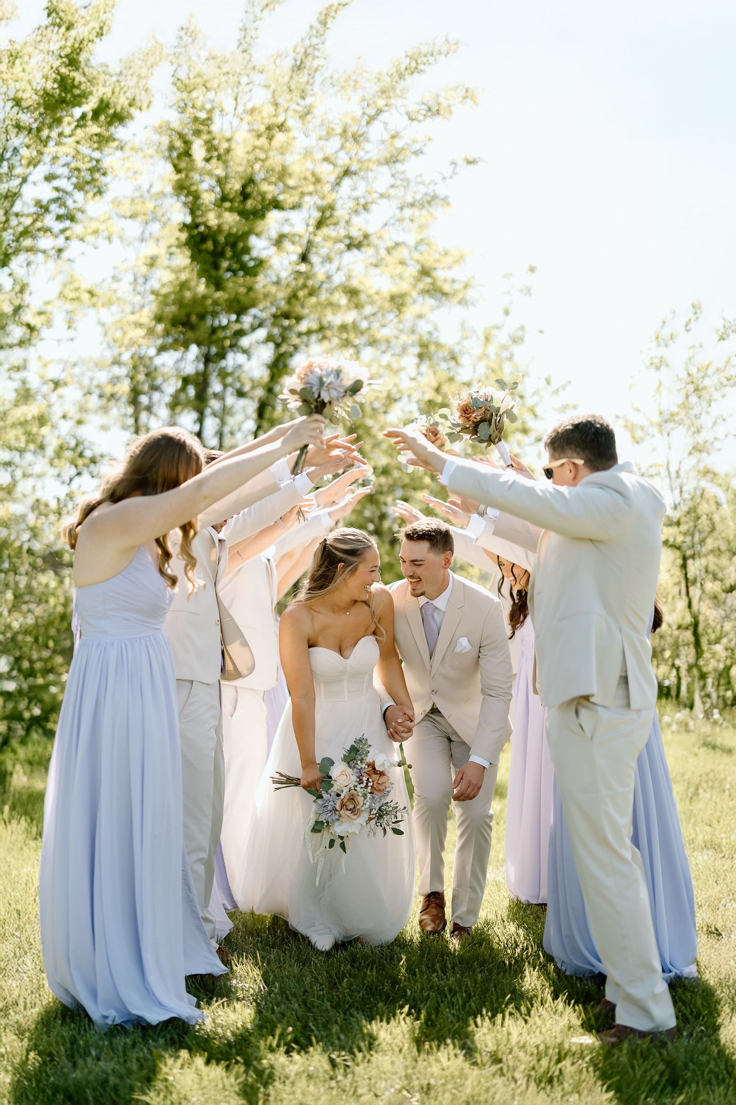 Wedding party forms tunnel with raised arms as couple runs underneath during outdoor photo session.