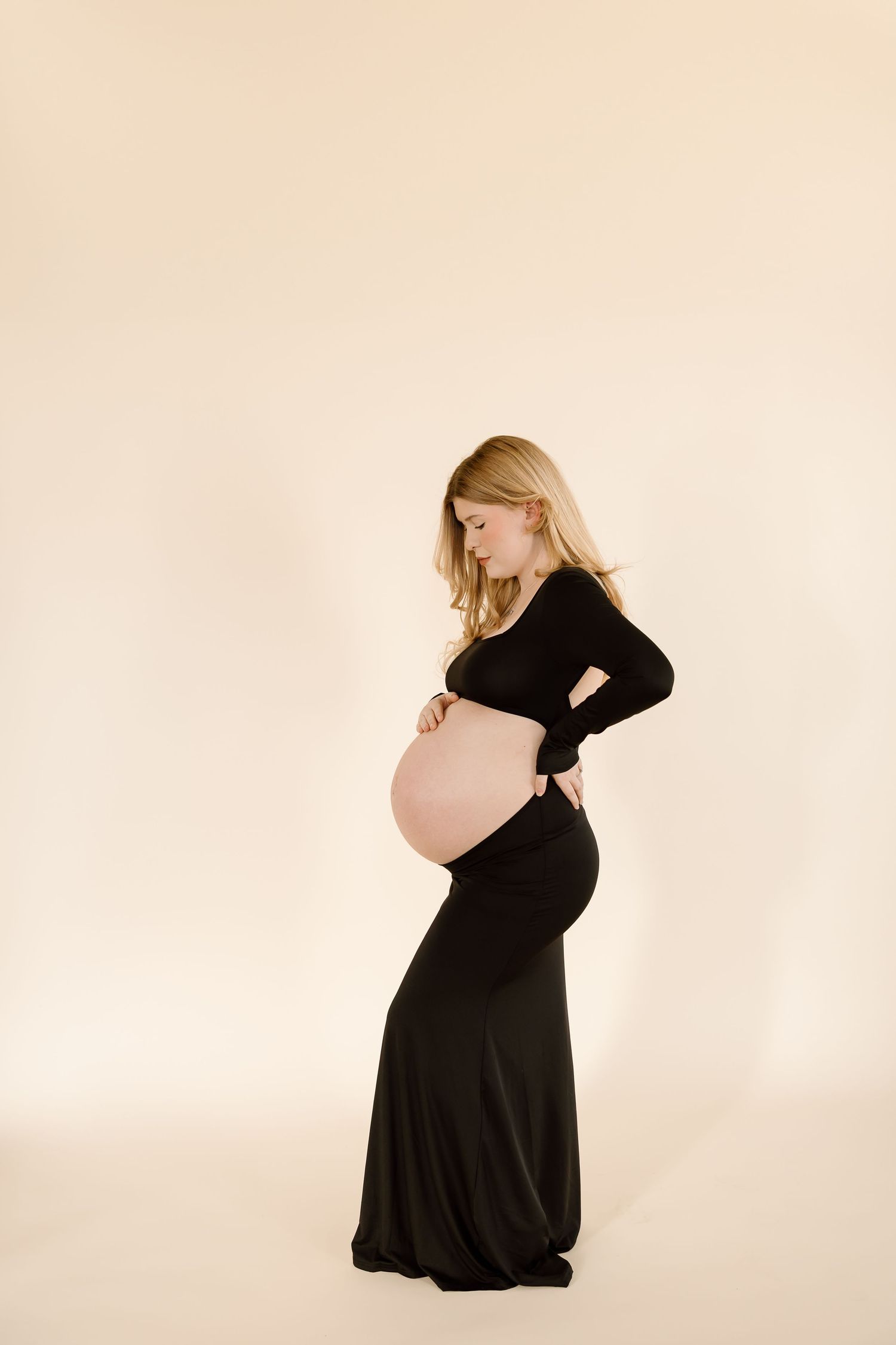 Full length maternity photo featuring elegant black dress and neutral studio background.