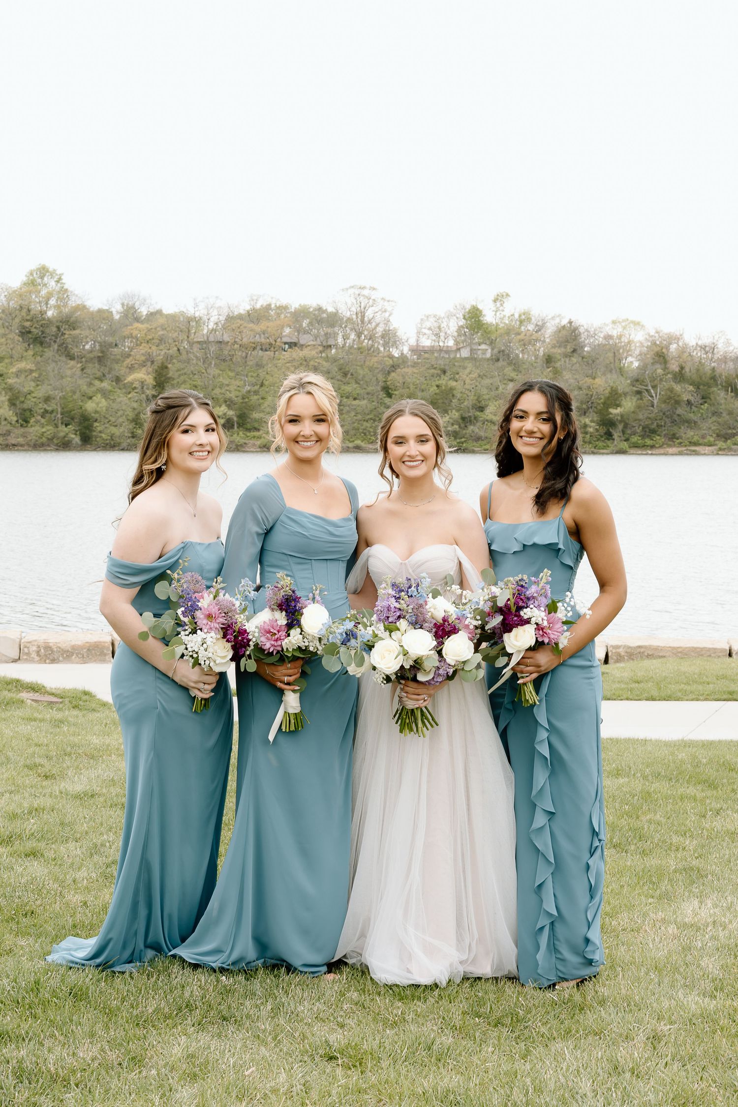 Four women in elegant blue gowns pose with bouquets by a lake.