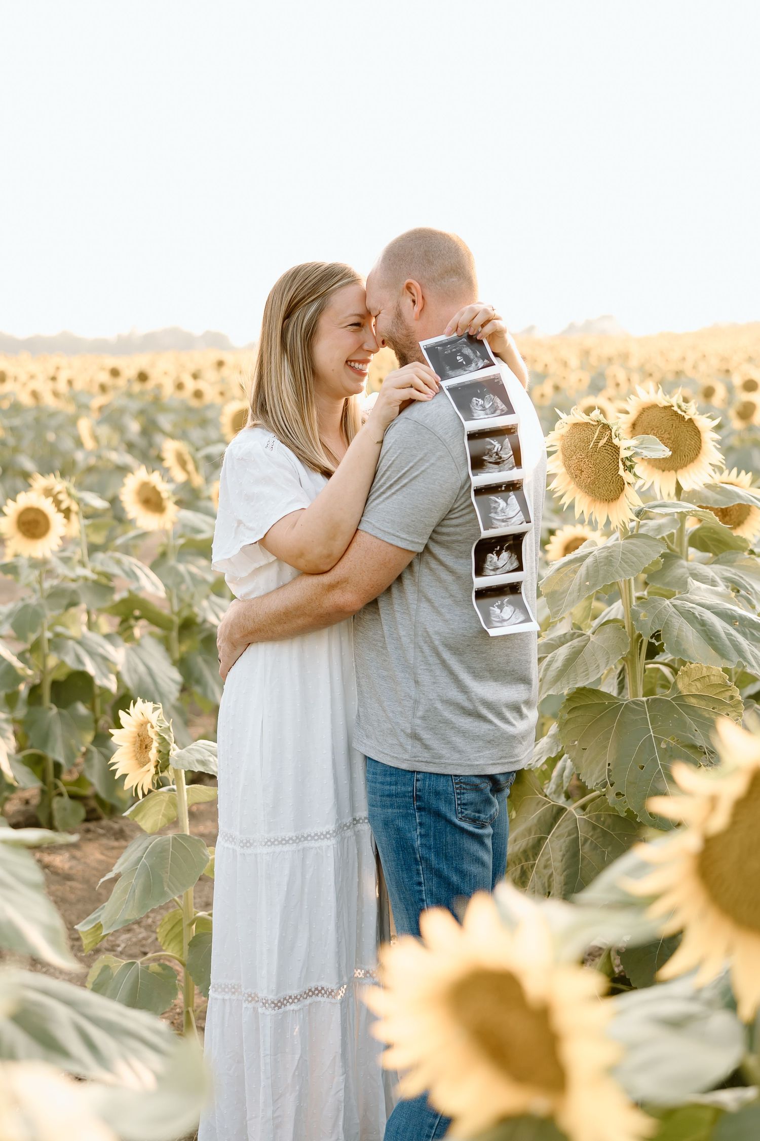 Couple sharing intimate moment among blooming sunflowers while holding ultrasound photos at sunset.