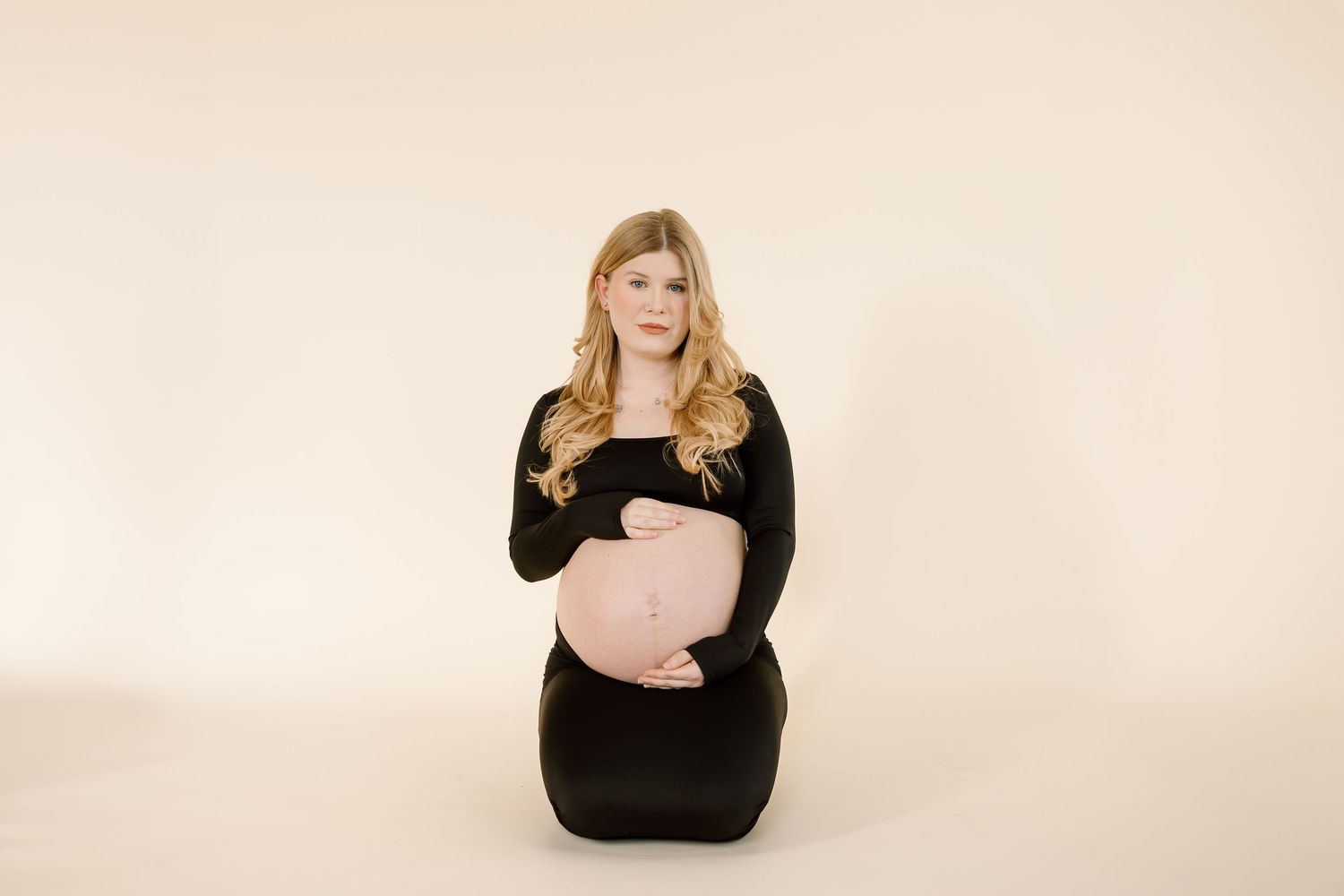 A pregnant woman wearing a black dress poses on her knees against a beige backdrop during a maternity photoshoot.