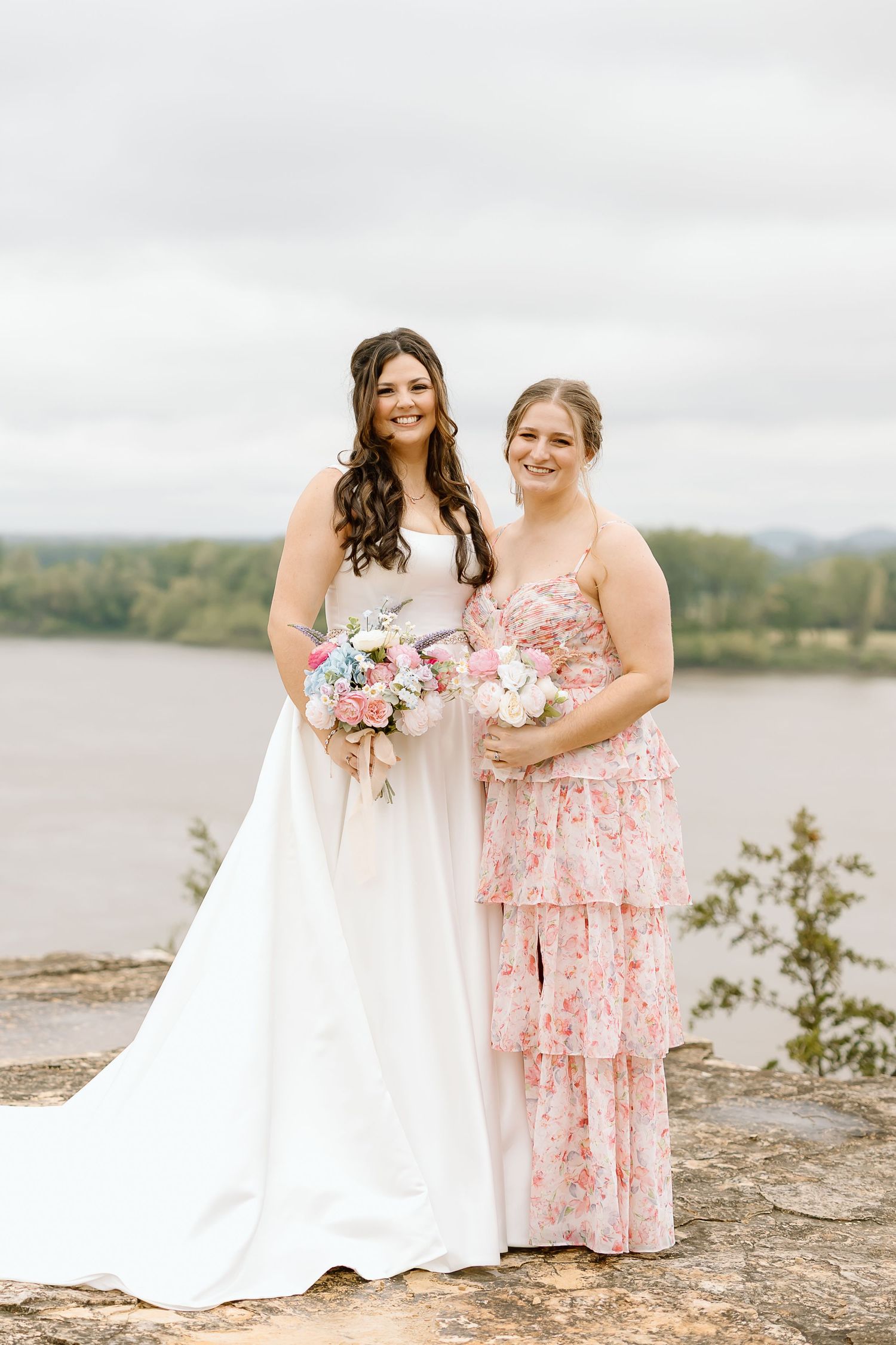 Two people in pink floral dresses pose together on rocky terrain near a lake during a wedding photo session.
