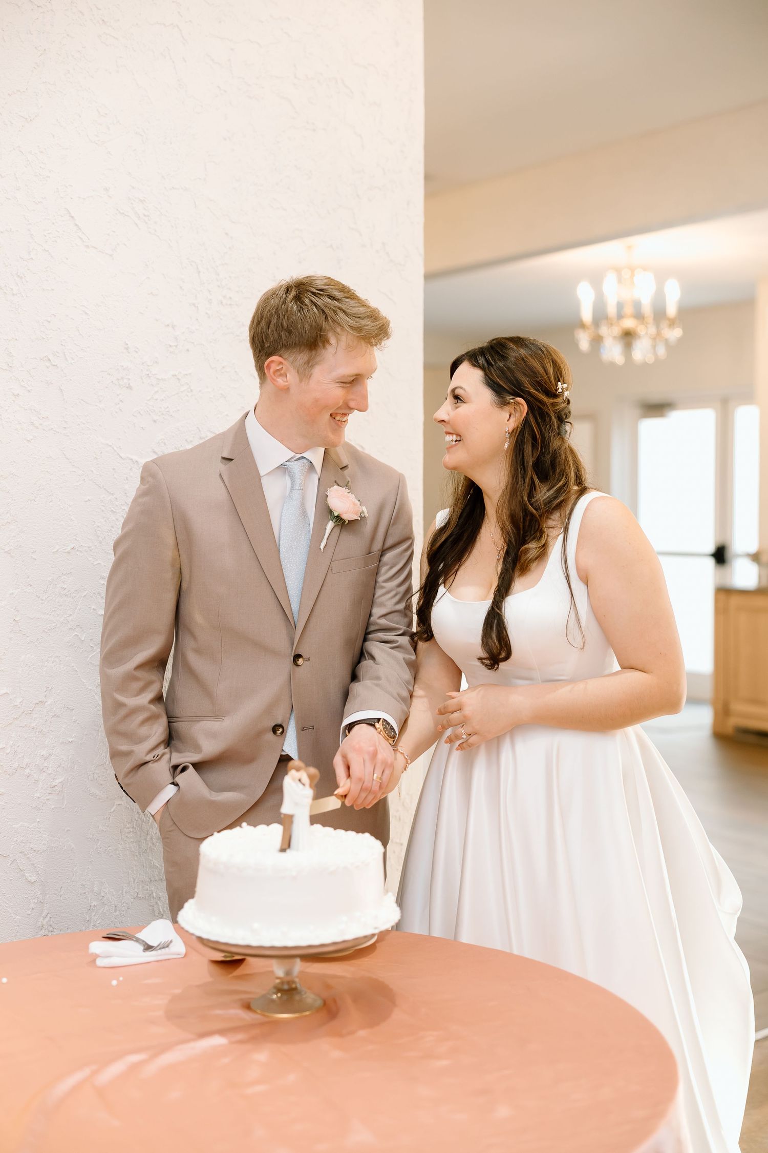 A couple in wedding attire share a joyful moment while cutting their simple white wedding cake at a reception.