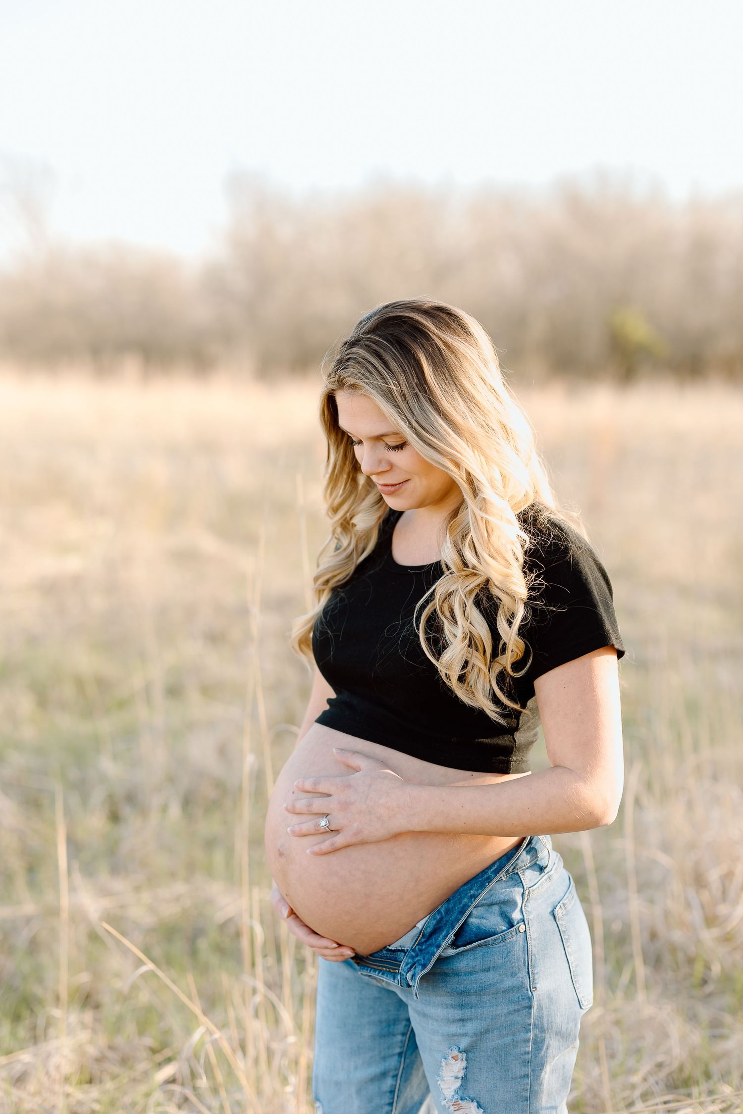 A pregnant woman in jeans and black shirt cradles her baby bump while standing in a sunlit field.