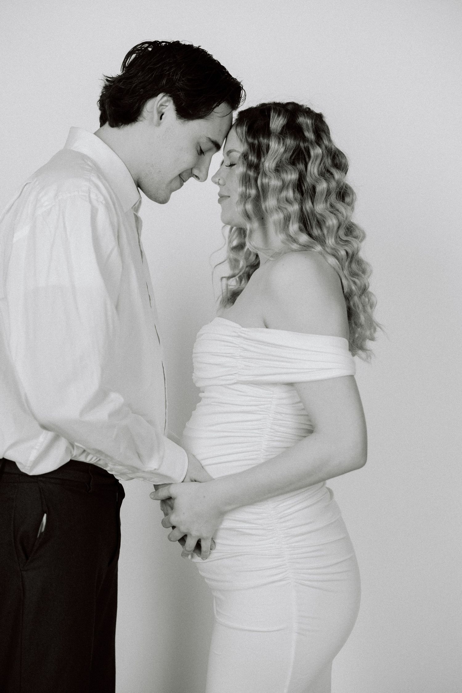 A tender black and white maternity photo showing a couple in intimate embrace with the woman wearing a fitted white dress.
