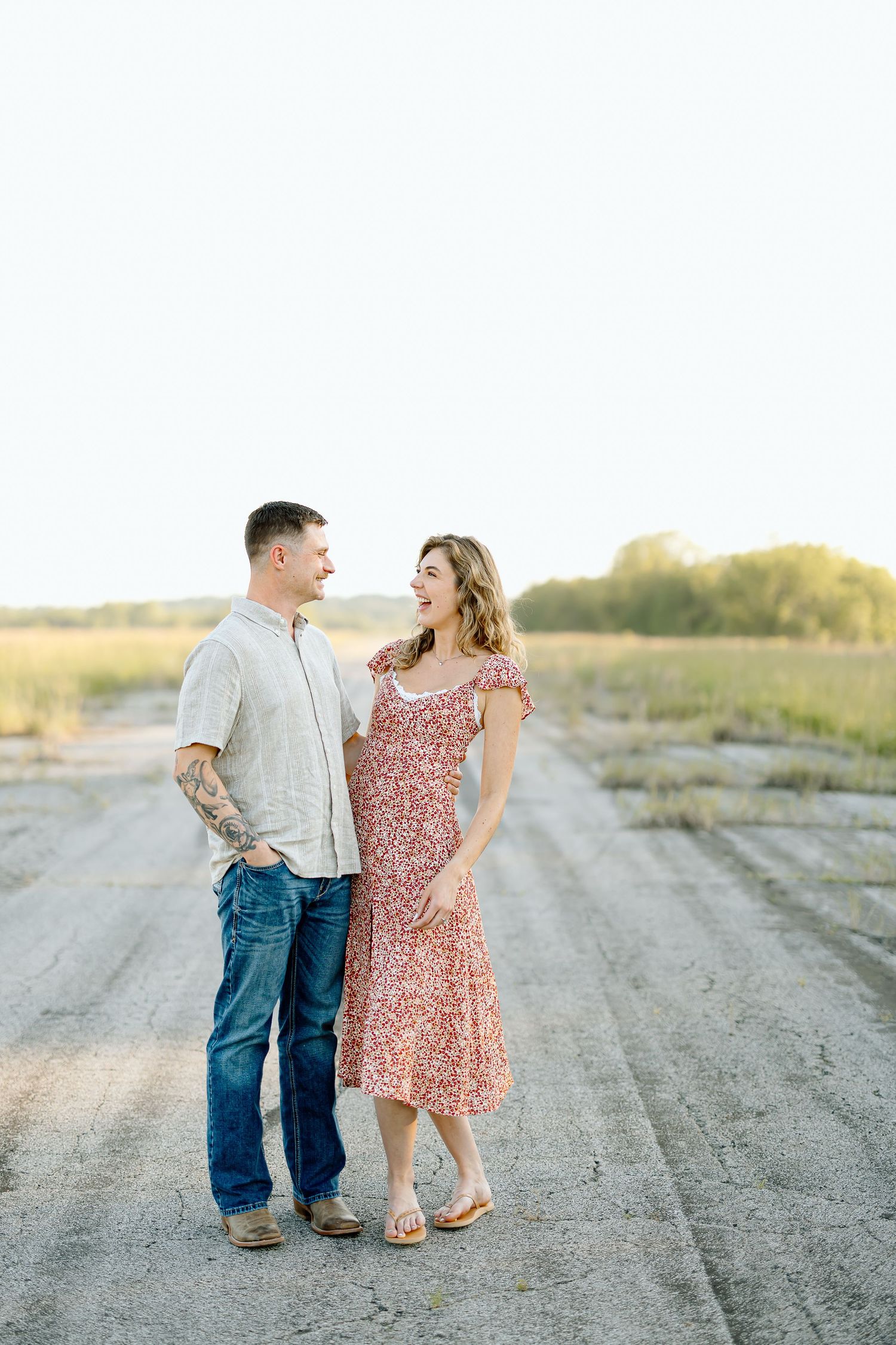 A couple in casual attire stands together on a dirt road at sunset in a romantic countryside setting.