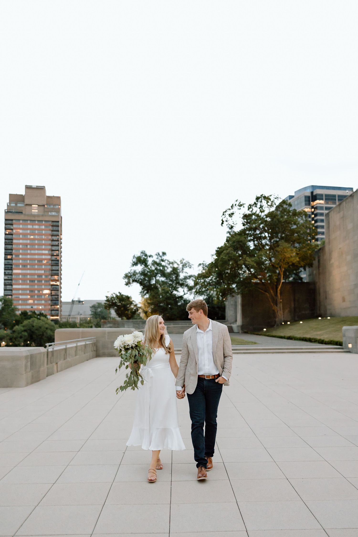 A couple in wedding attire walks away together on a concrete rooftop terrace with city buildings in the background.