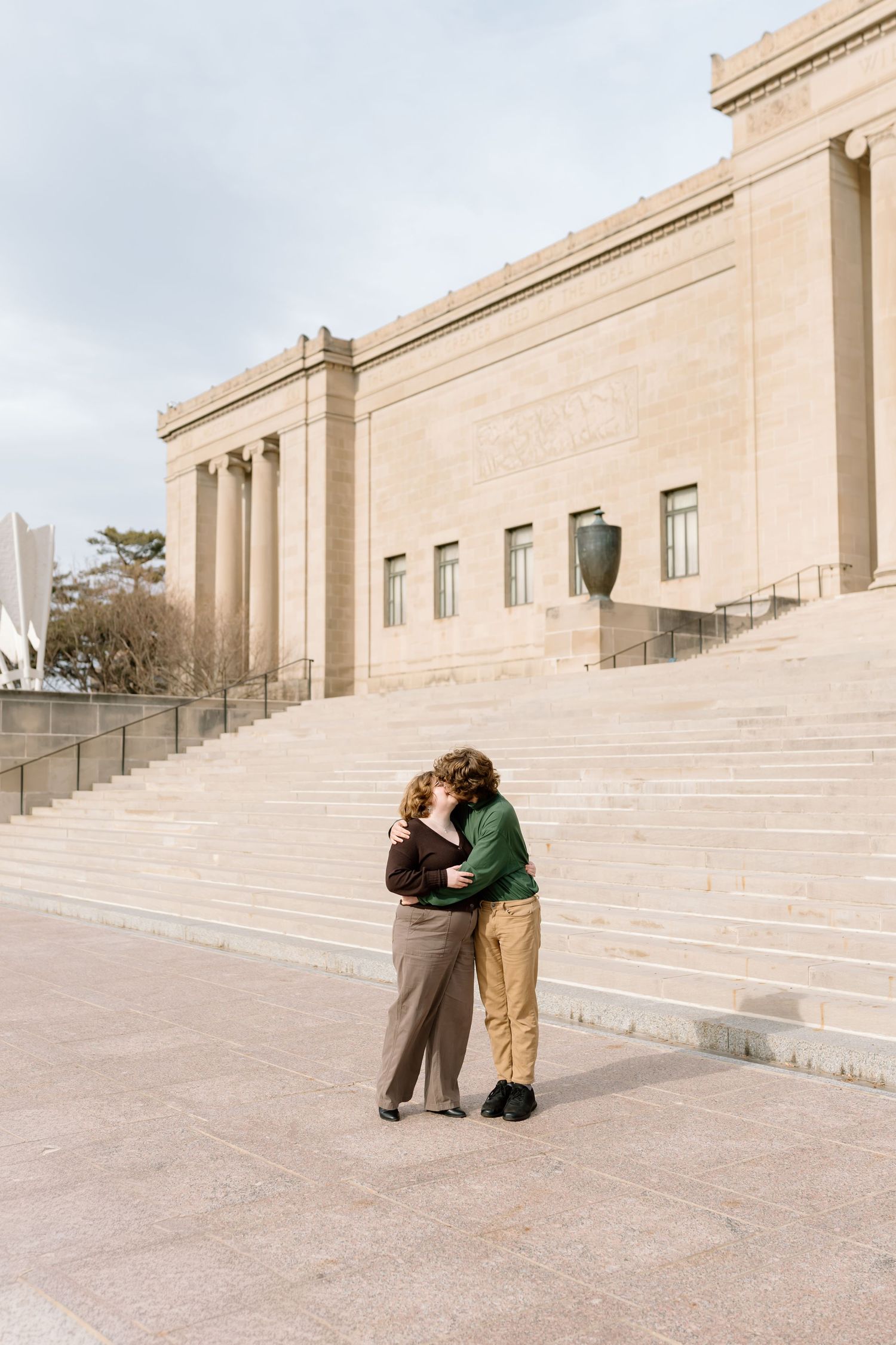 Two people embrace on wide stone steps of a grand building with limestone facade and columns.
