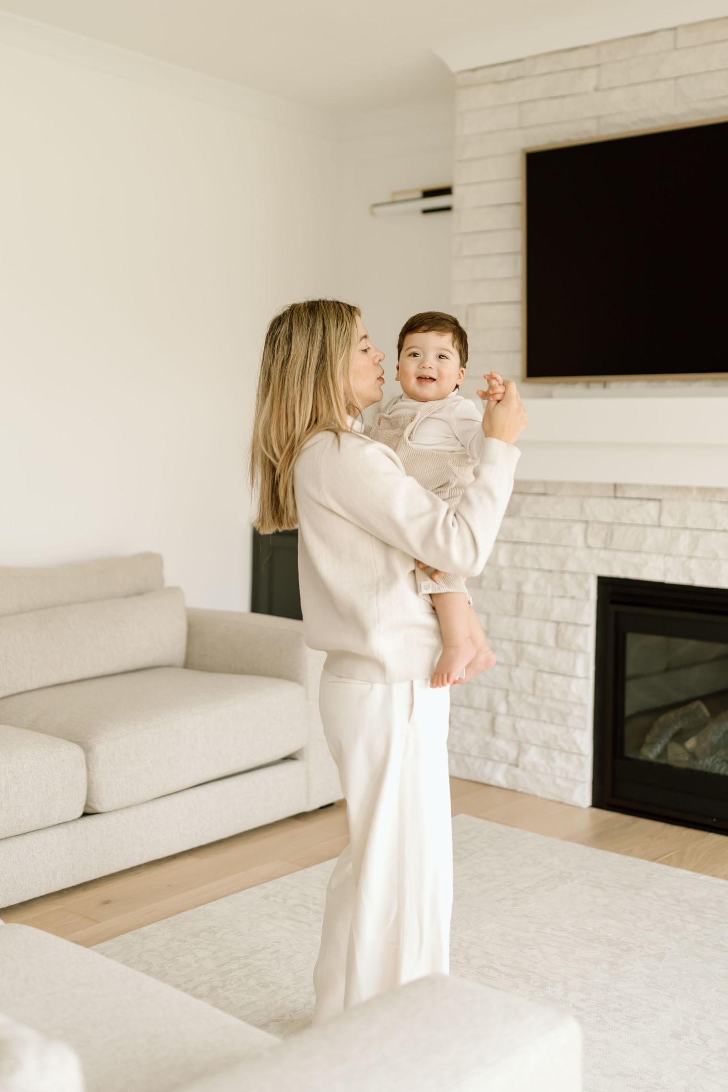 A parent and baby share a joyful moment in a bright modern living room with white decor and a fireplace.