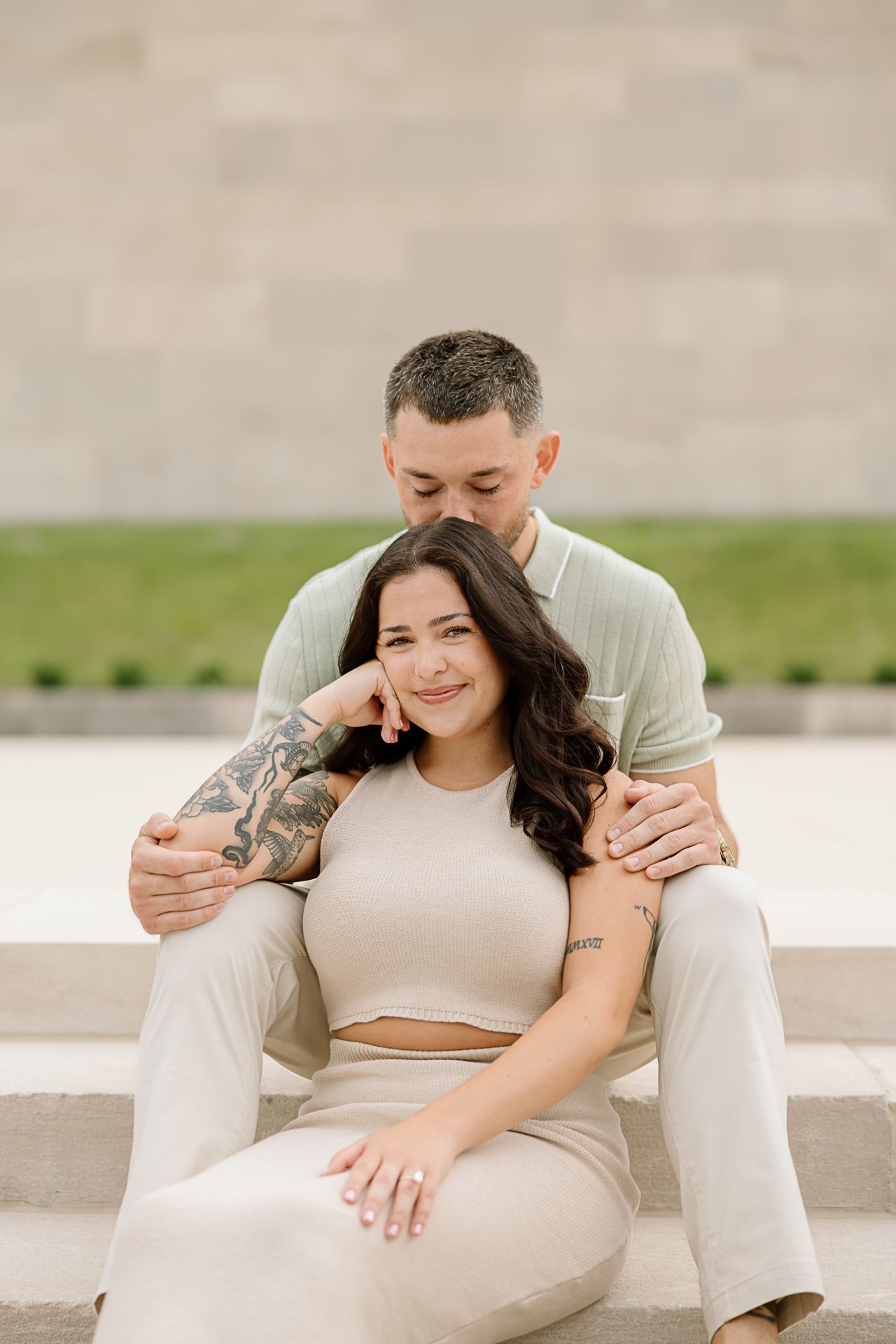 A couple in light colored clothing shares an intimate moment while sitting on concrete steps outdoors.