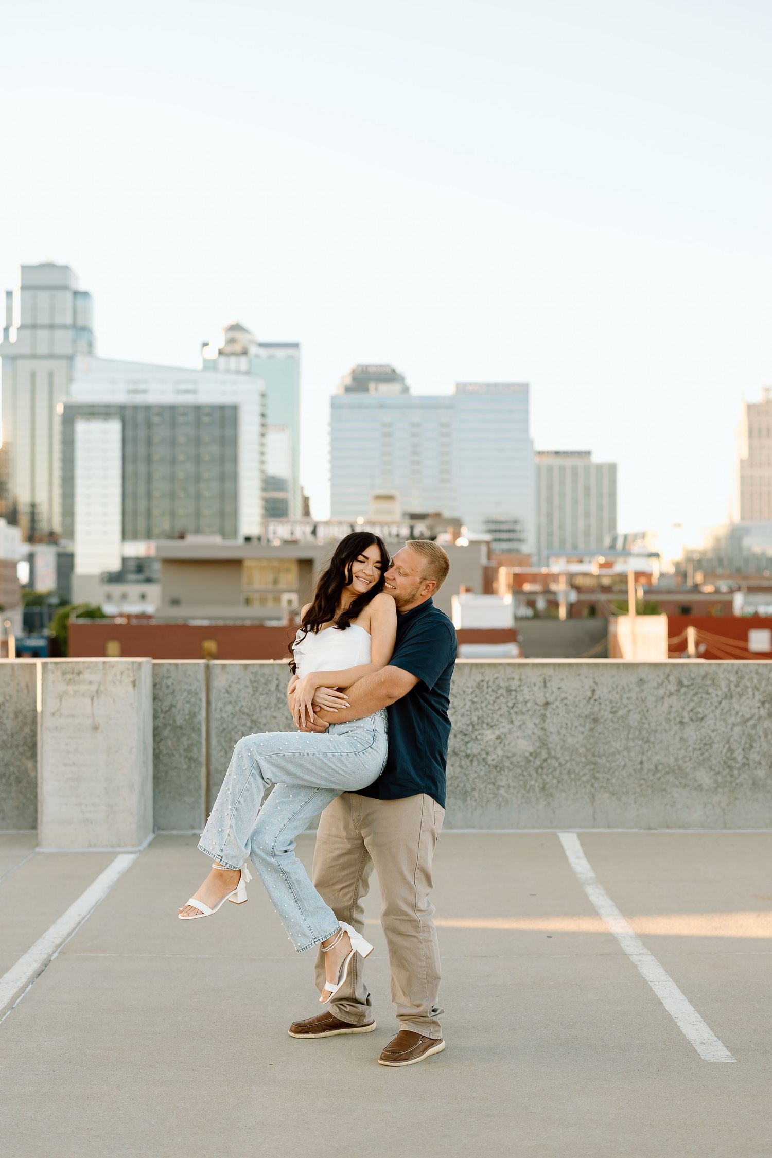 A romantic couple dances together on a rooftop parking garage with a city skyline in the background.