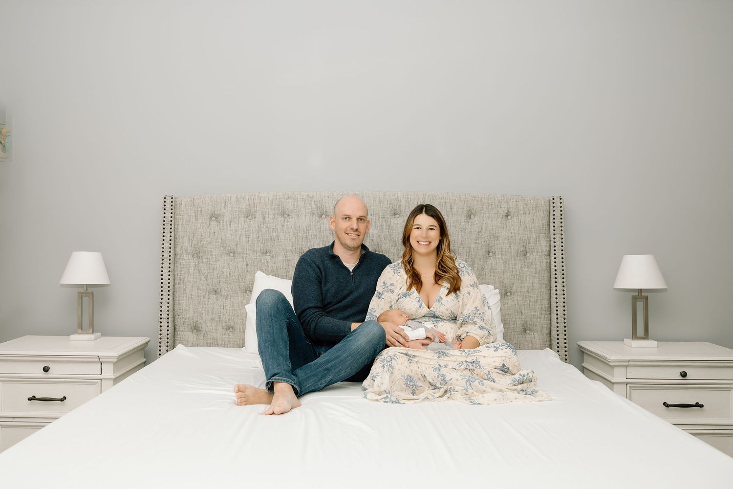A couple sits together on a gray tufted headboard bed with matching white nightstands and table lamps on either side.
