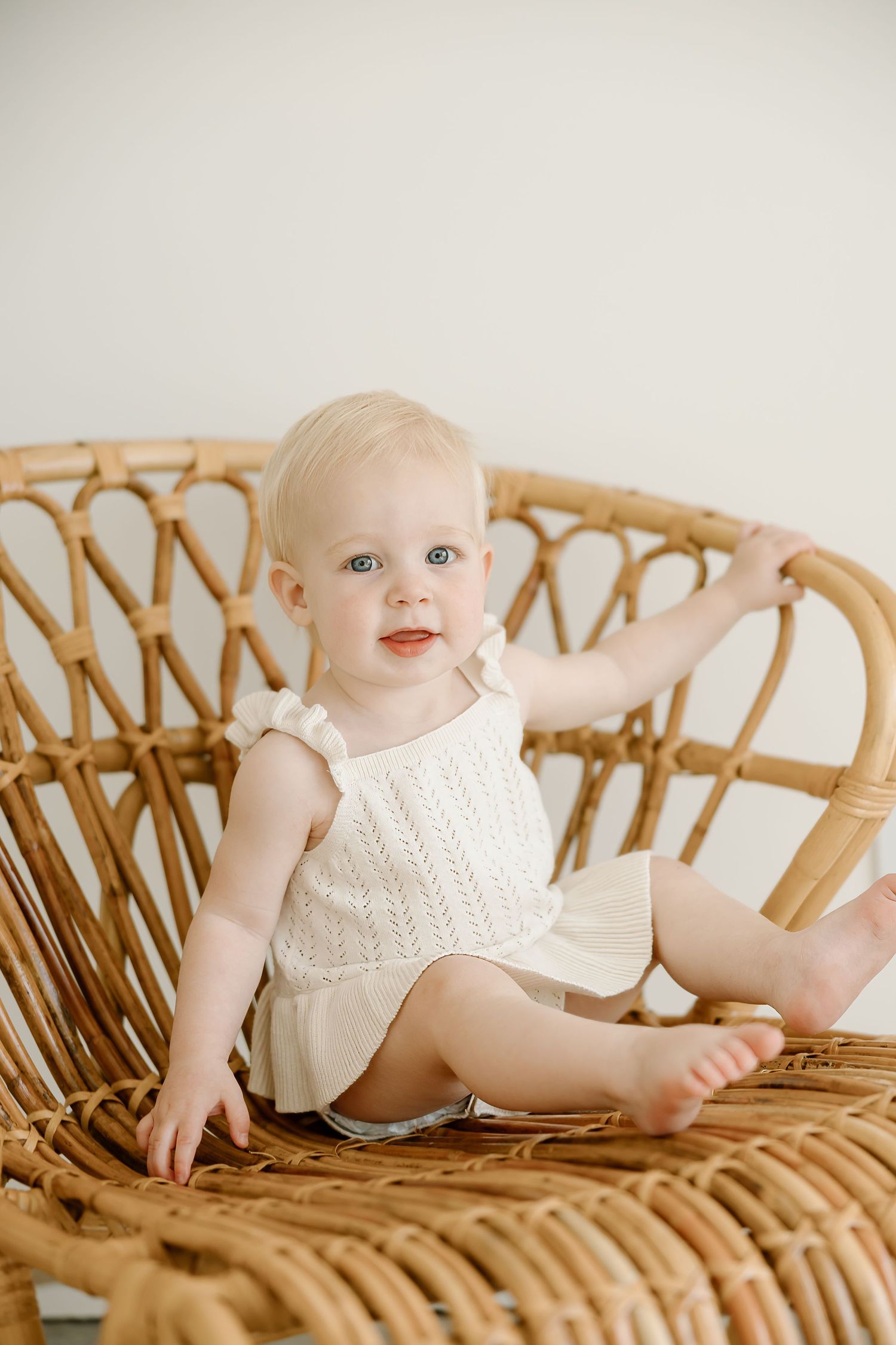 Baby in white knit outfit sits in vintage rattan peacock chair against neutral background.