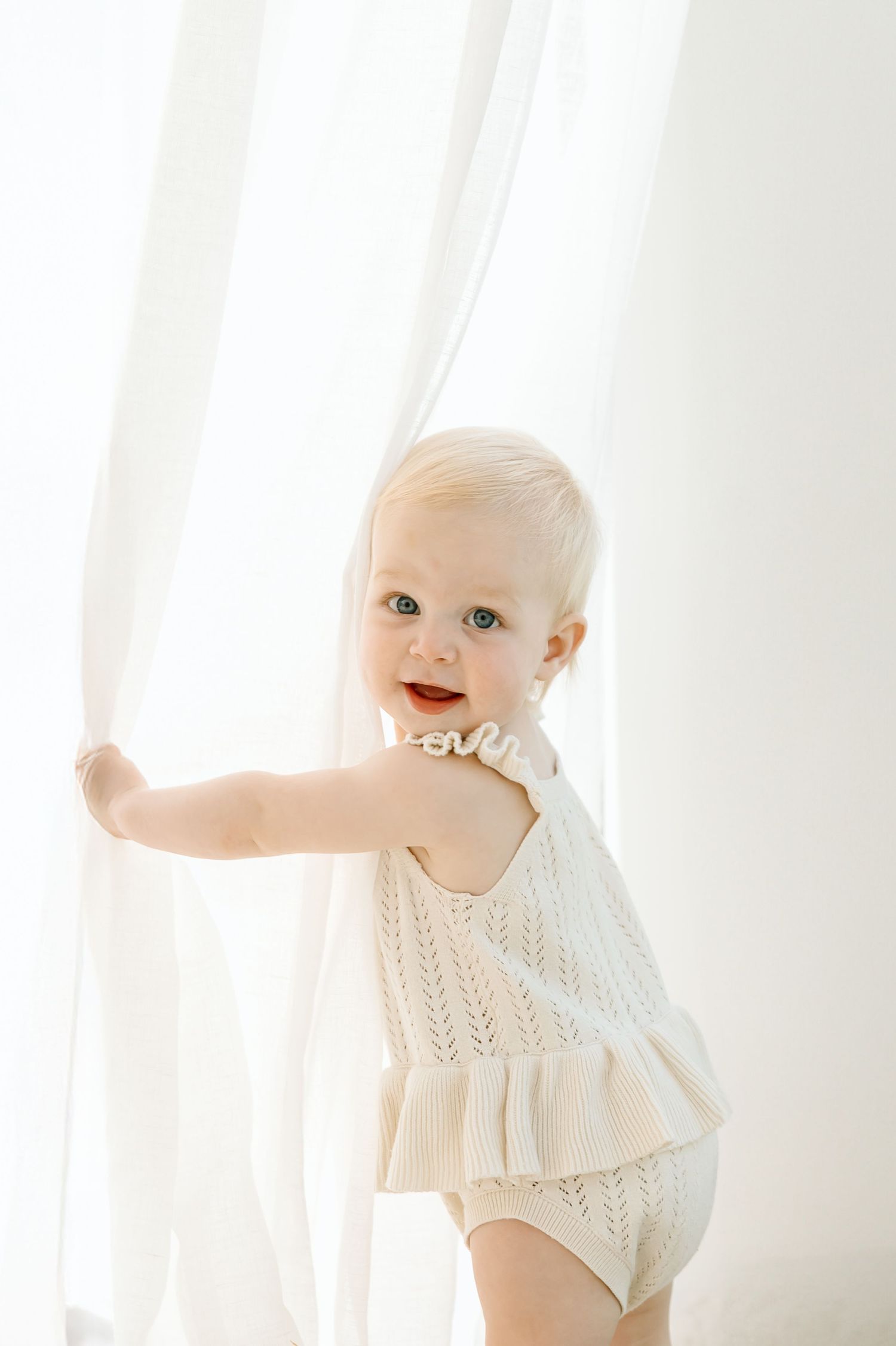 Baby in white knit sweater smiles while playing with sheer curtains in bright sunlit room.