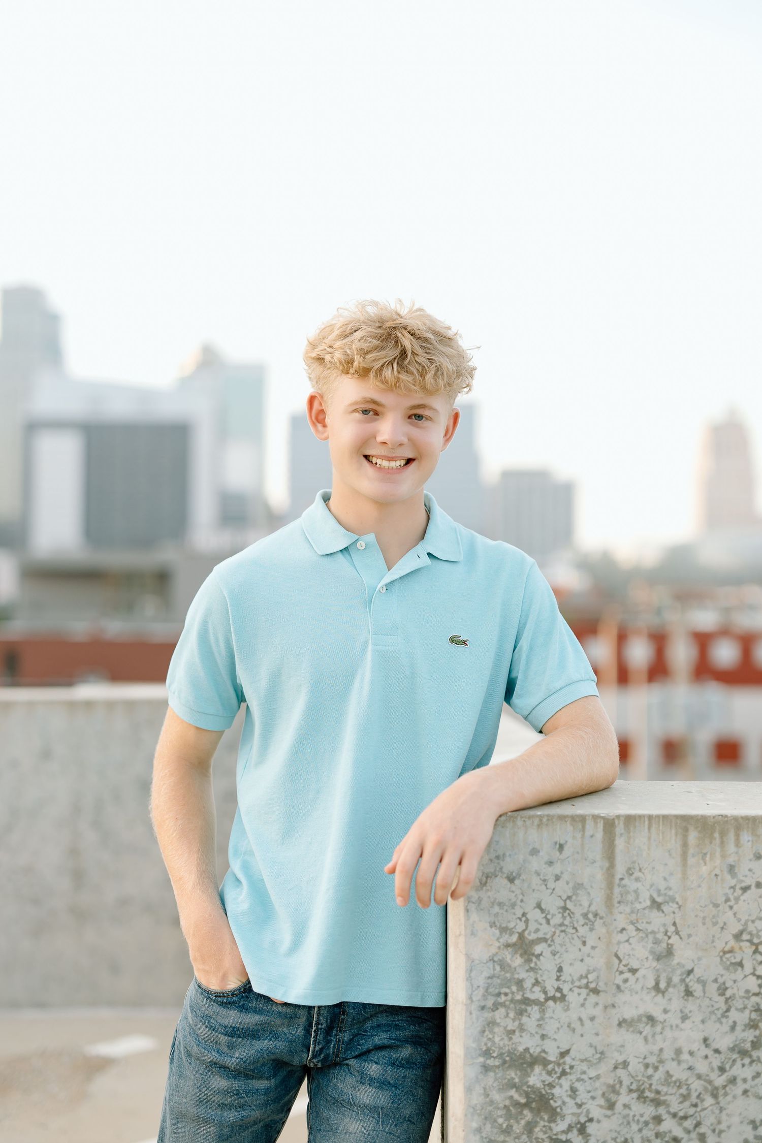 Young person in light blue polo shirt smiling on rooftop with city skyline in background.