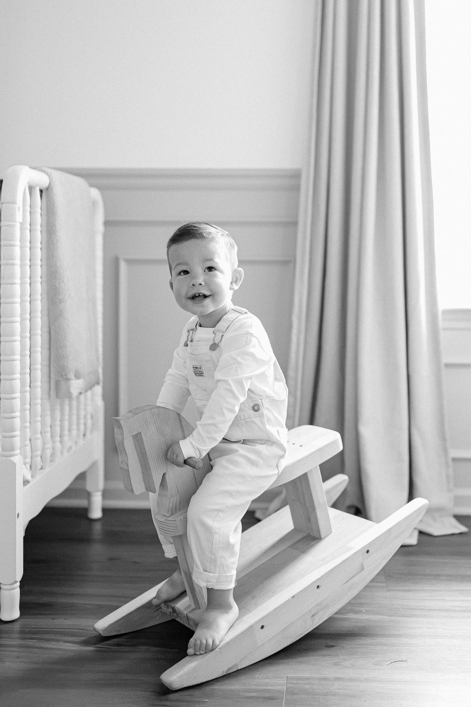A baby in white clothing sits and plays on a wooden rocking toy in a bright minimalist room.