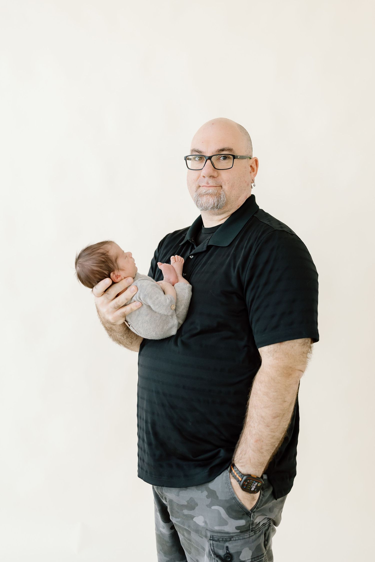 Someone in a black polo shirt holds a small baby while standing against a white background.