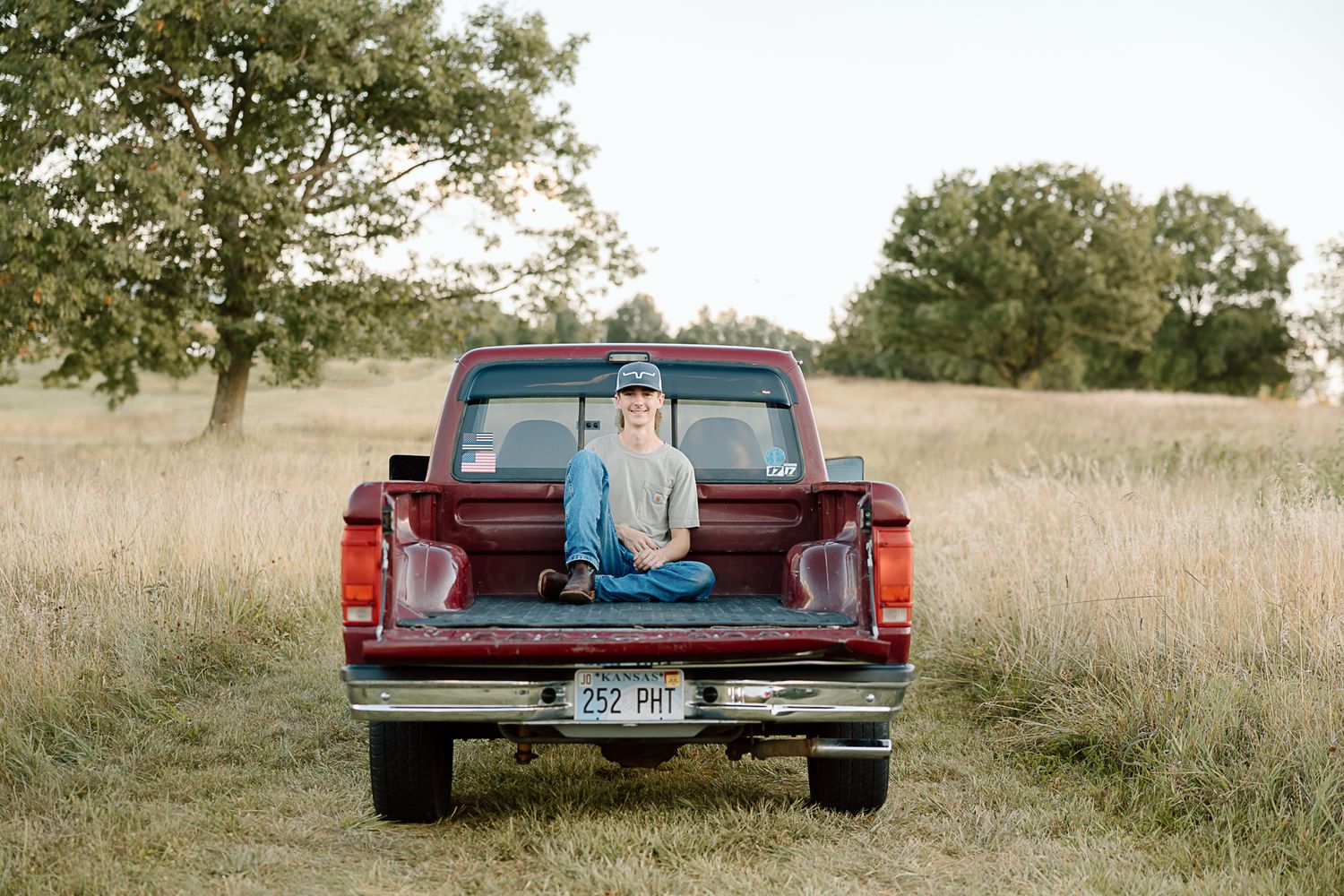 A vintage red pickup truck parked in a golden grass field during sunset with a romantic couple sitting in the truck bed.