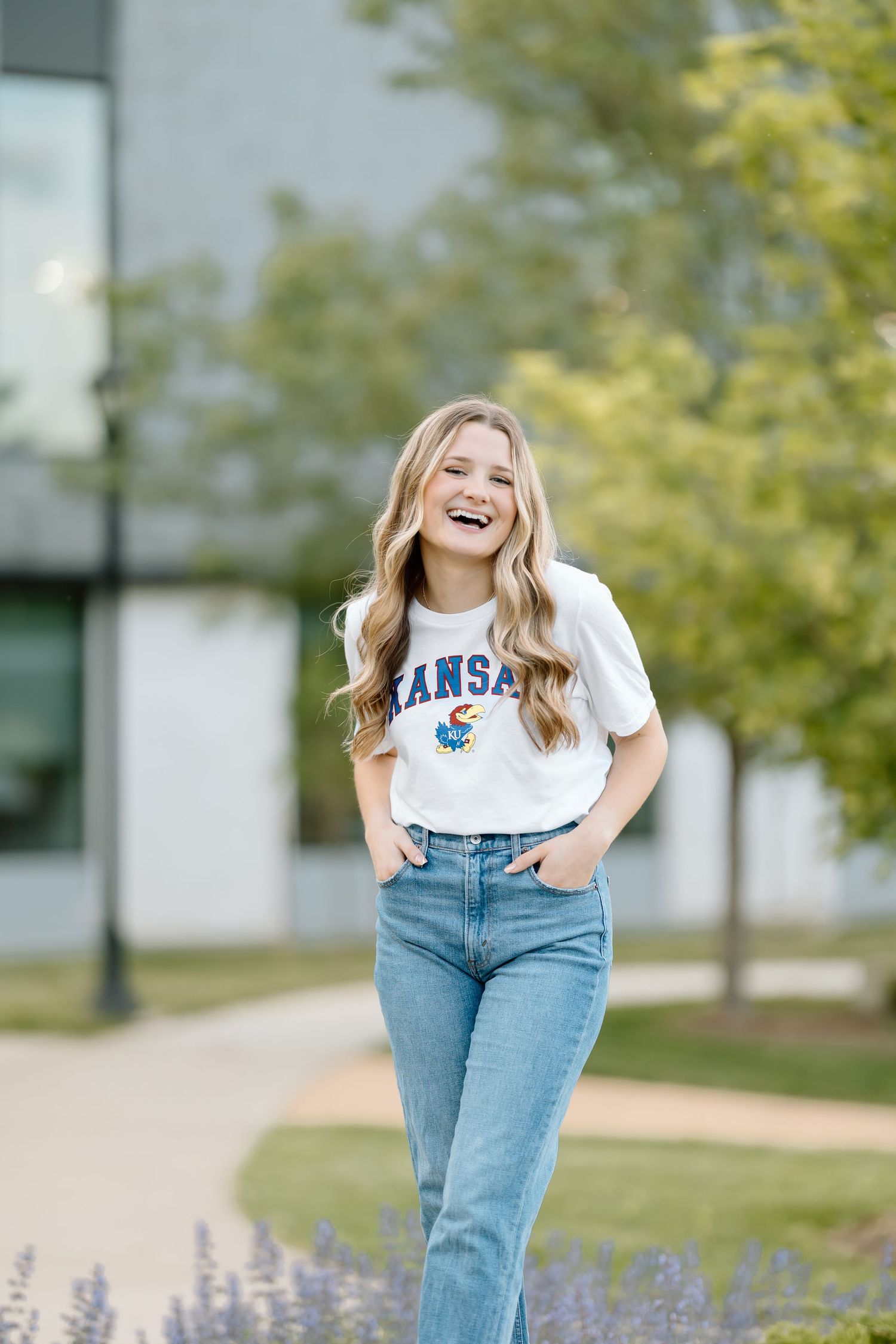 A person wearing a Kansas t-shirt and jeans stands on a pathway surrounded by greenery, laughing joyfully.