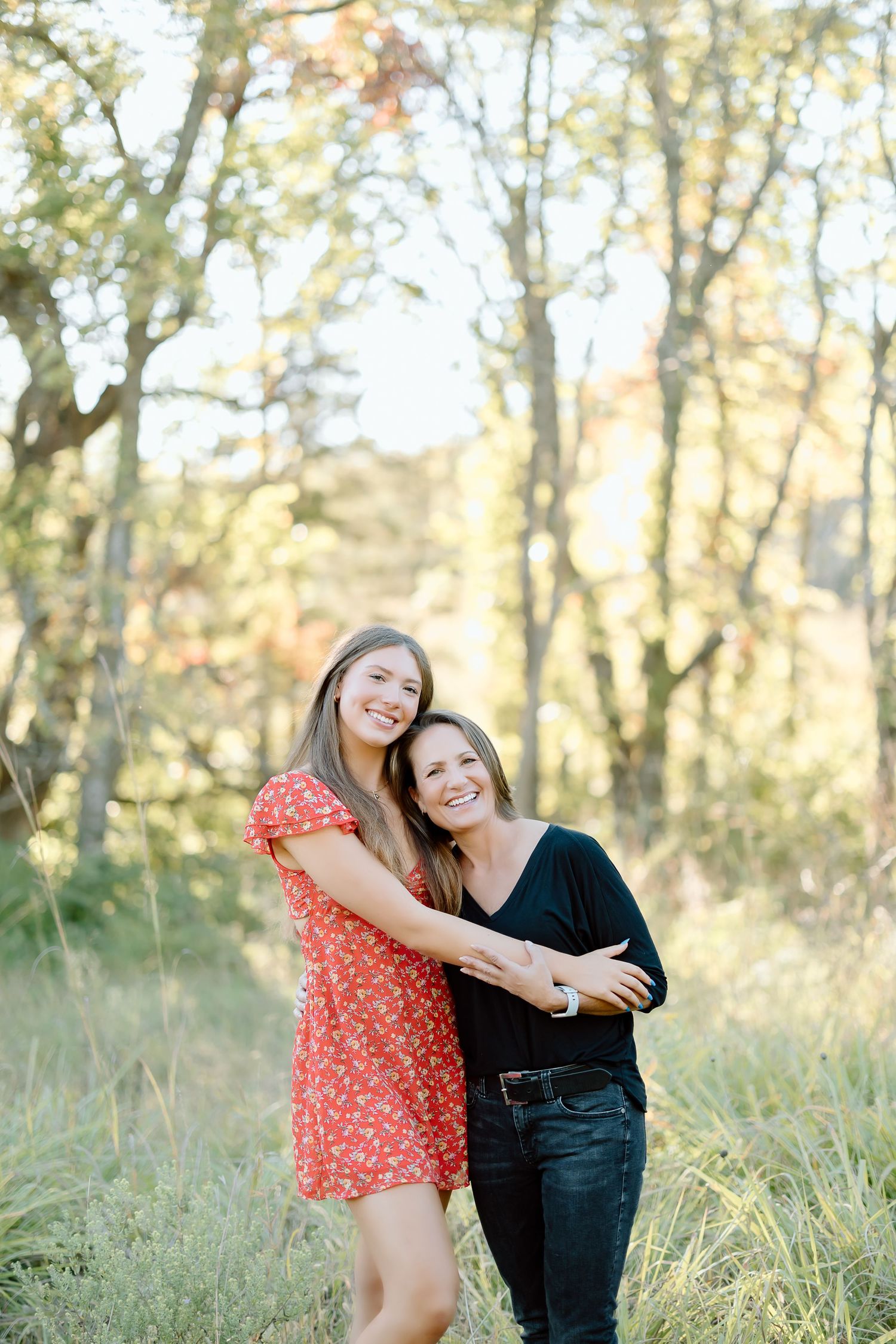 Two friends share a joyful moment together in a sun-dappled forest setting with tall trees and golden light.