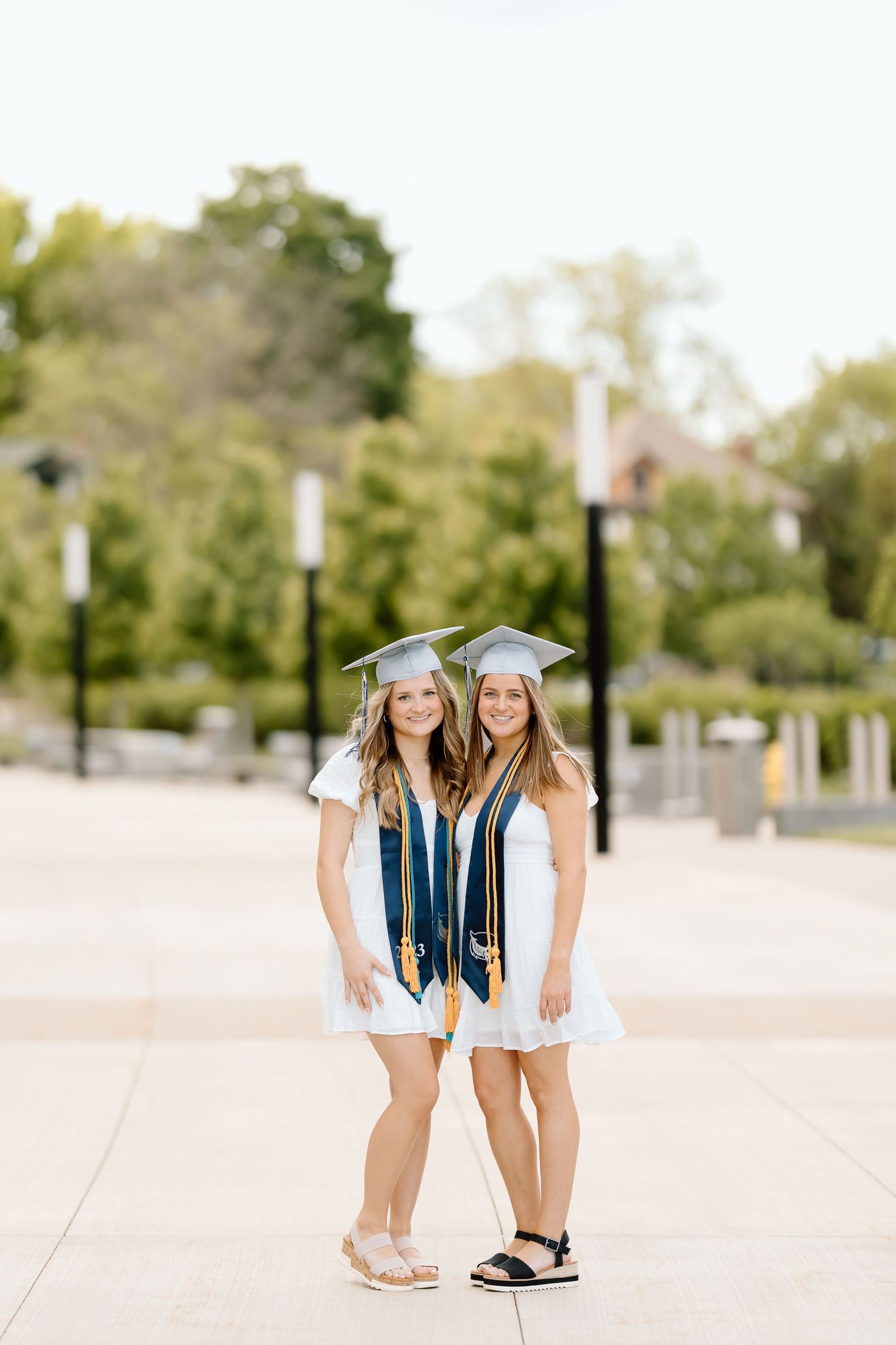 Two graduates in white dresses and graduation caps stand together in a sunny outdoor plaza.