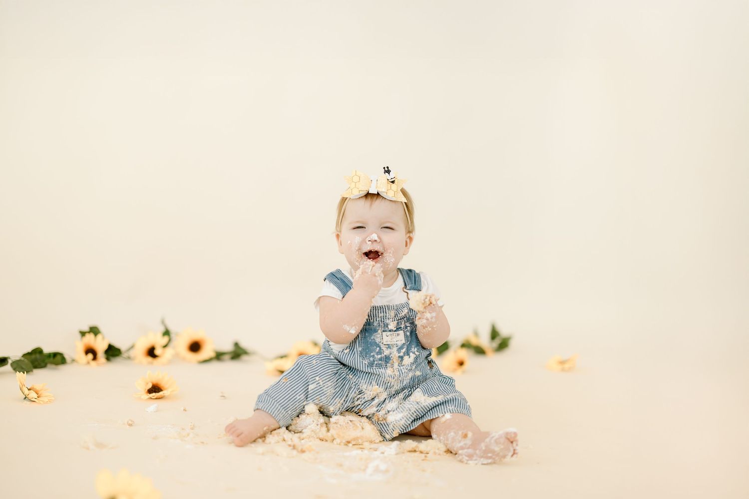 Baby in light blue outfit sits on white backdrop surrounded by scattered autumn leaves and pumpkins.
