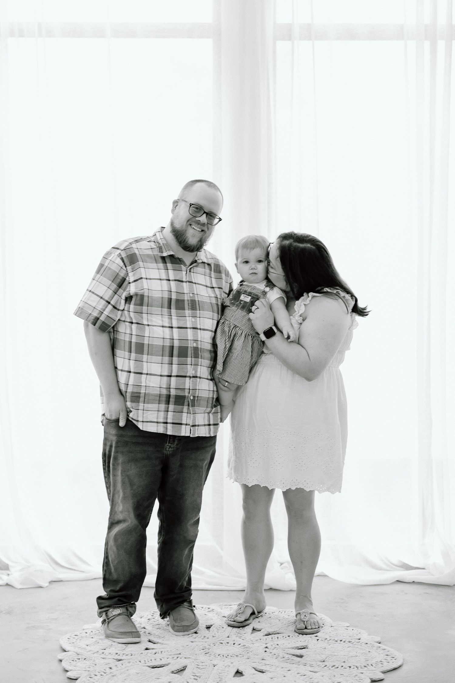 Black and white family portrait with parents and small child standing together against bright window backdrop.